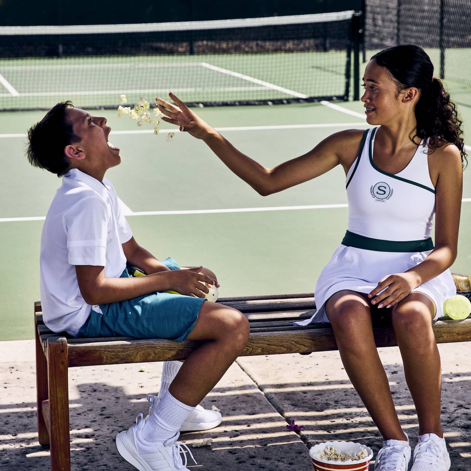 On a shaded wooden bench in front of a pickleball court, two children enjoy some popcorn, pickleballs strewn on the ground.
