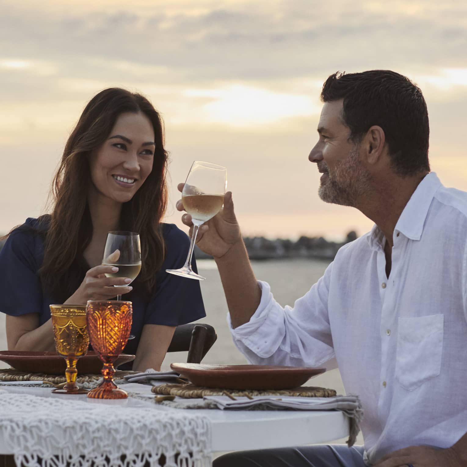 A smiling couple drinking wine against a sunset-streaked sky, at a beachfront dinner table set with amber-coloured goblets.