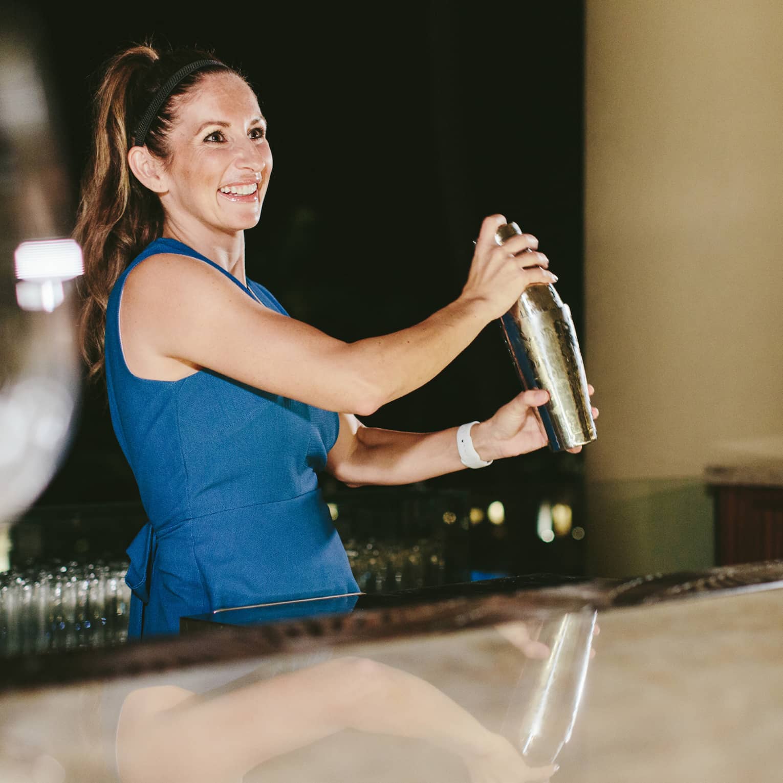 Smiling bartender in blue dress shaking a cocktail shaker behind a bar at night
