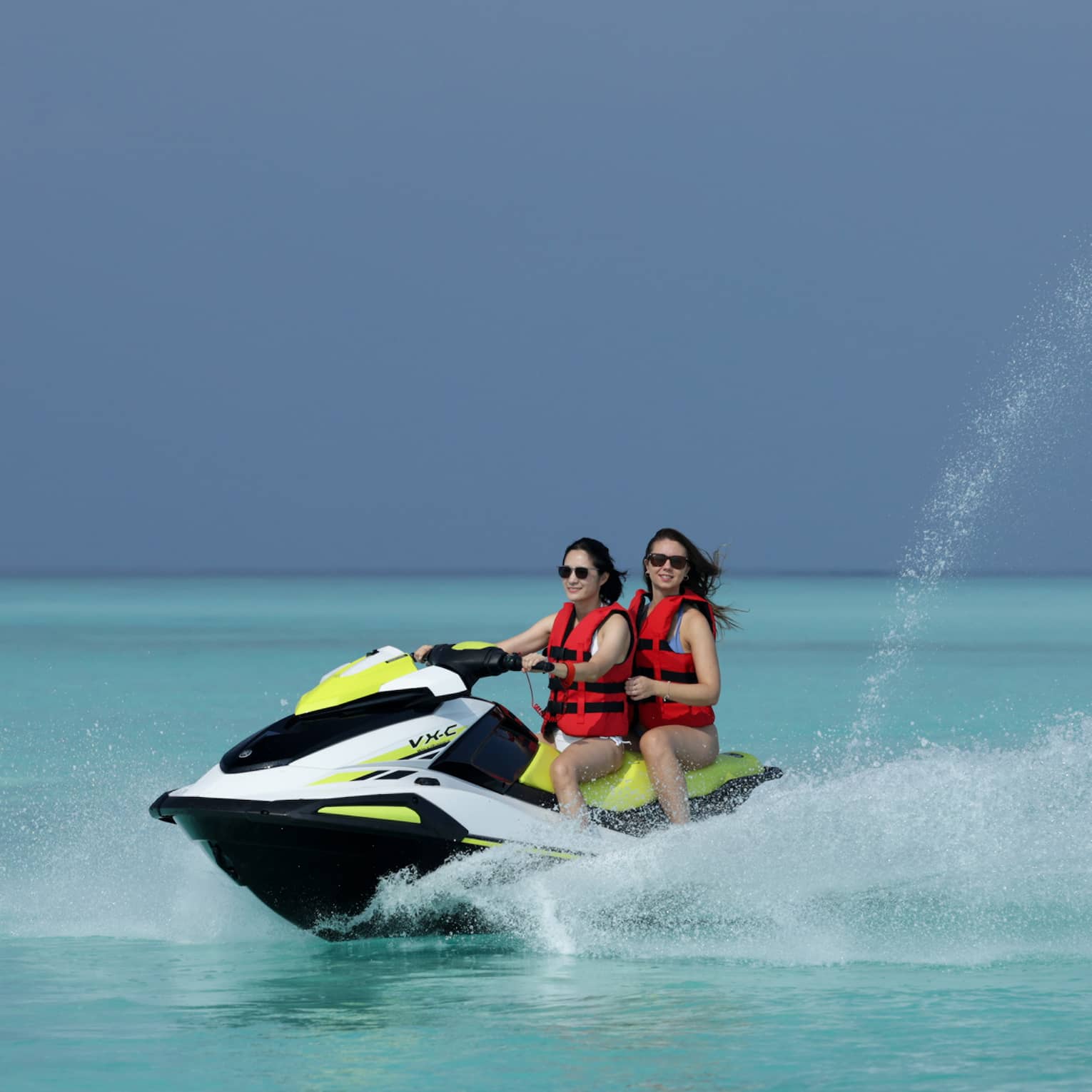 Wearing life jackets, two smiling jet-skiers ride through the clear ocean, water spurting up from beside and behind the boat.
