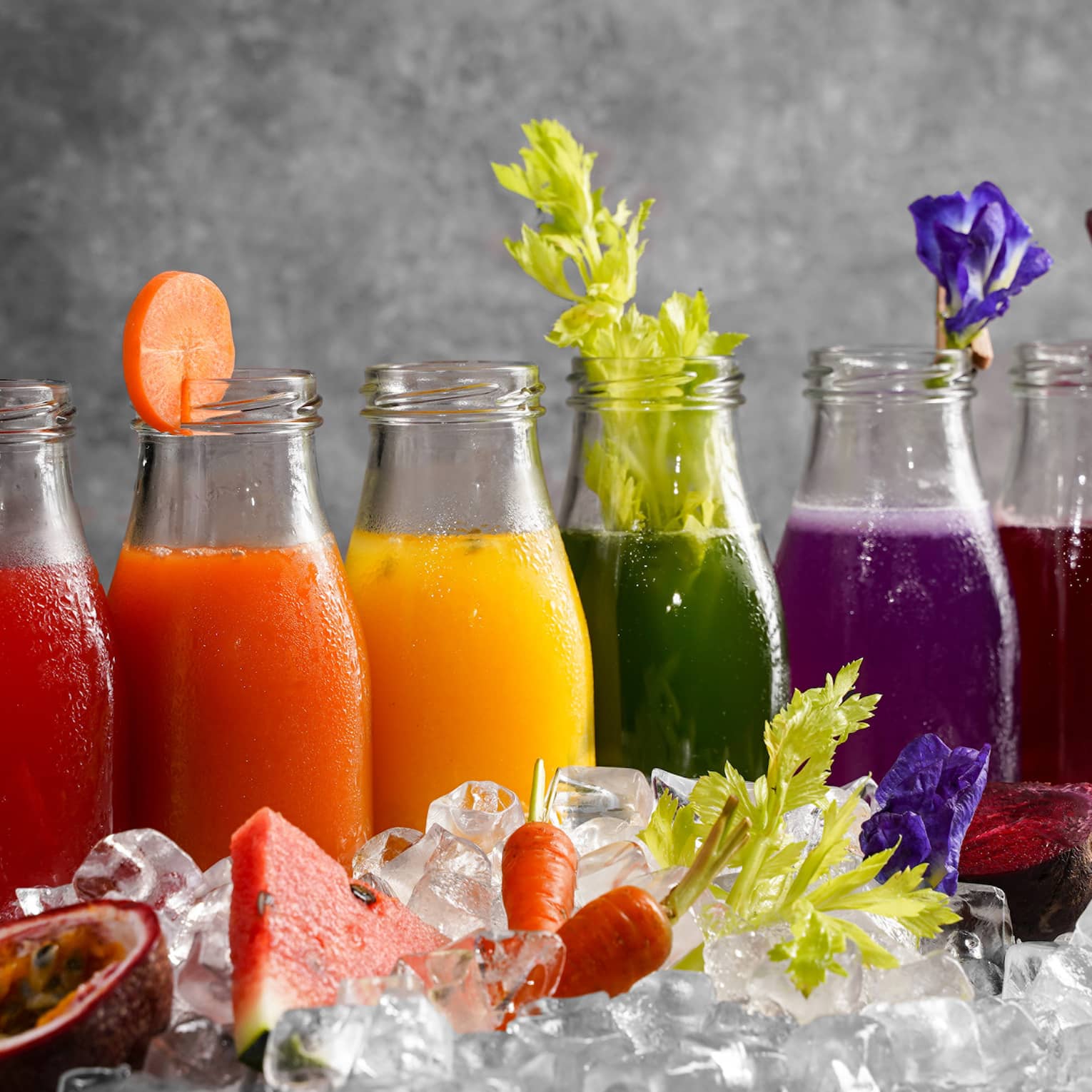 Six bottles of assorted colourful juices and garnishes lined up behind a pile of ice cubes