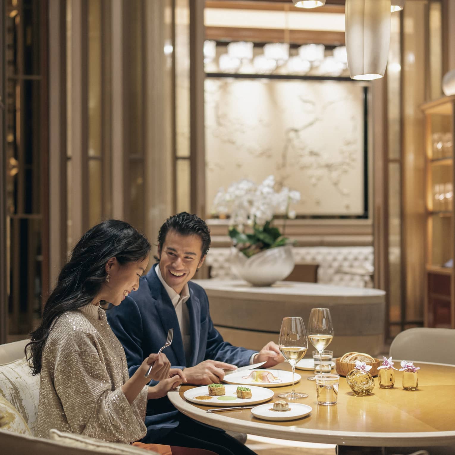A smiling couple savours lunch with wine in a circular booth at a fine dining restaurant with warm pendant lighting.