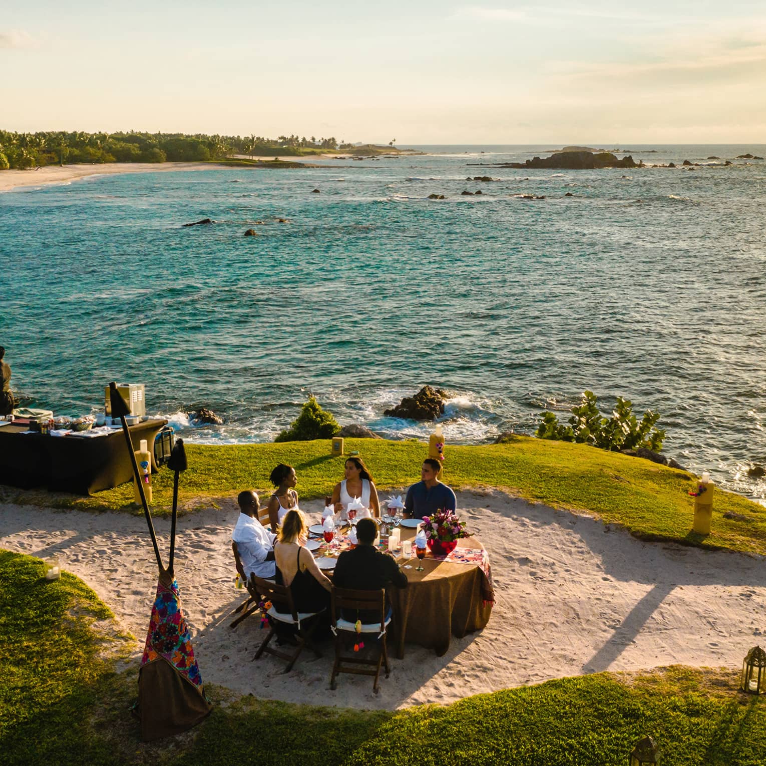 Aerial view of Dinner on the Rock dining table on lawn, sandy path near ocean at sunset