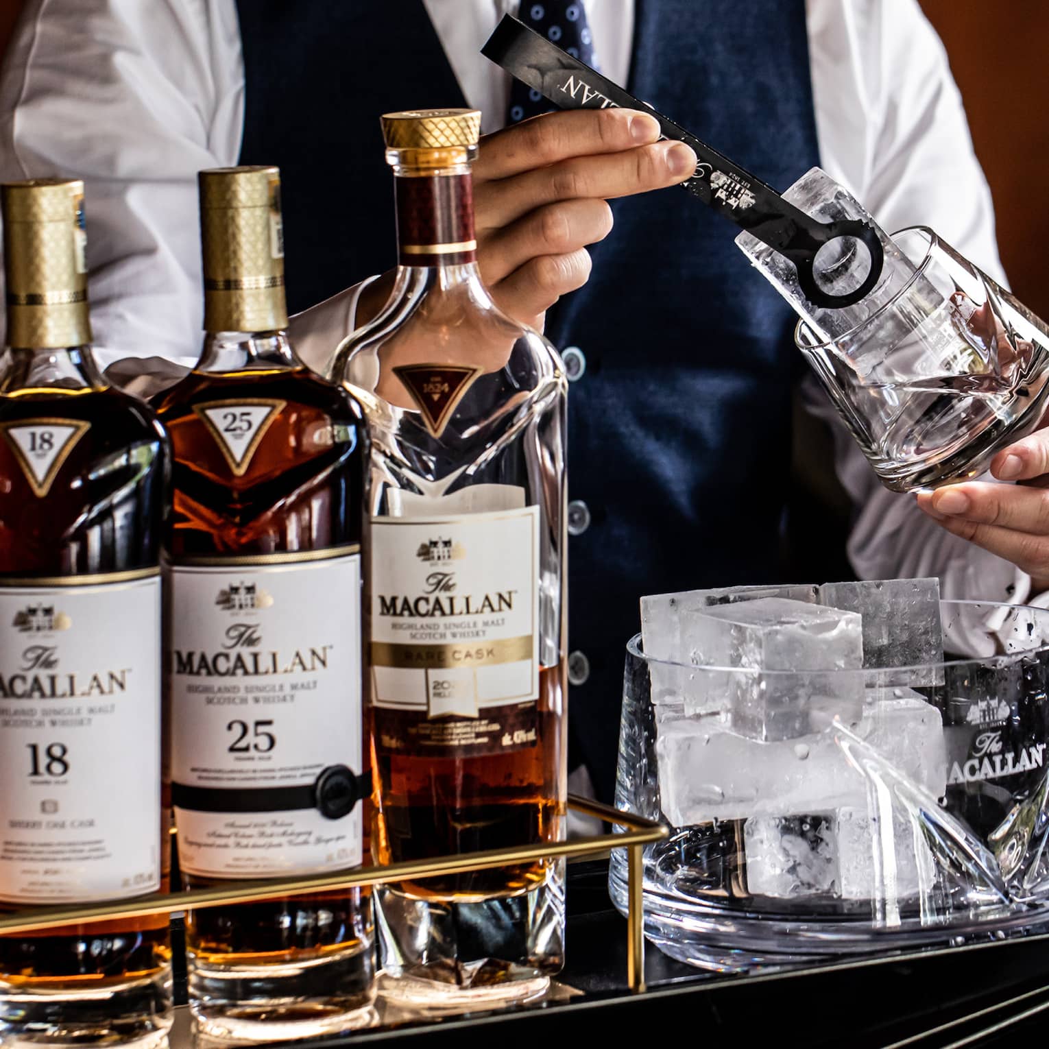 A bartender in tie and vest places a large ice cube in a glass with tongs, next to a five-bottle lineup of Macallan whiskies.