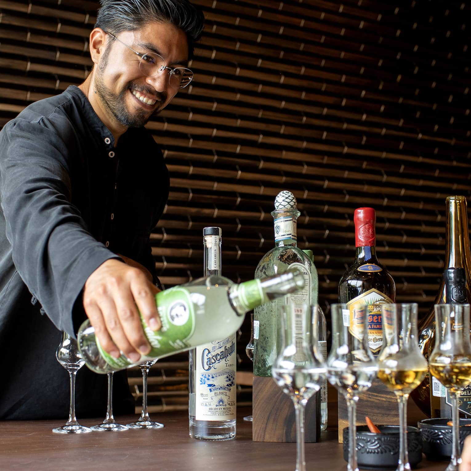 A bartender pours a clear liquor into grappa glasses, two others filled with an amber liquor, aside various other bottles.
