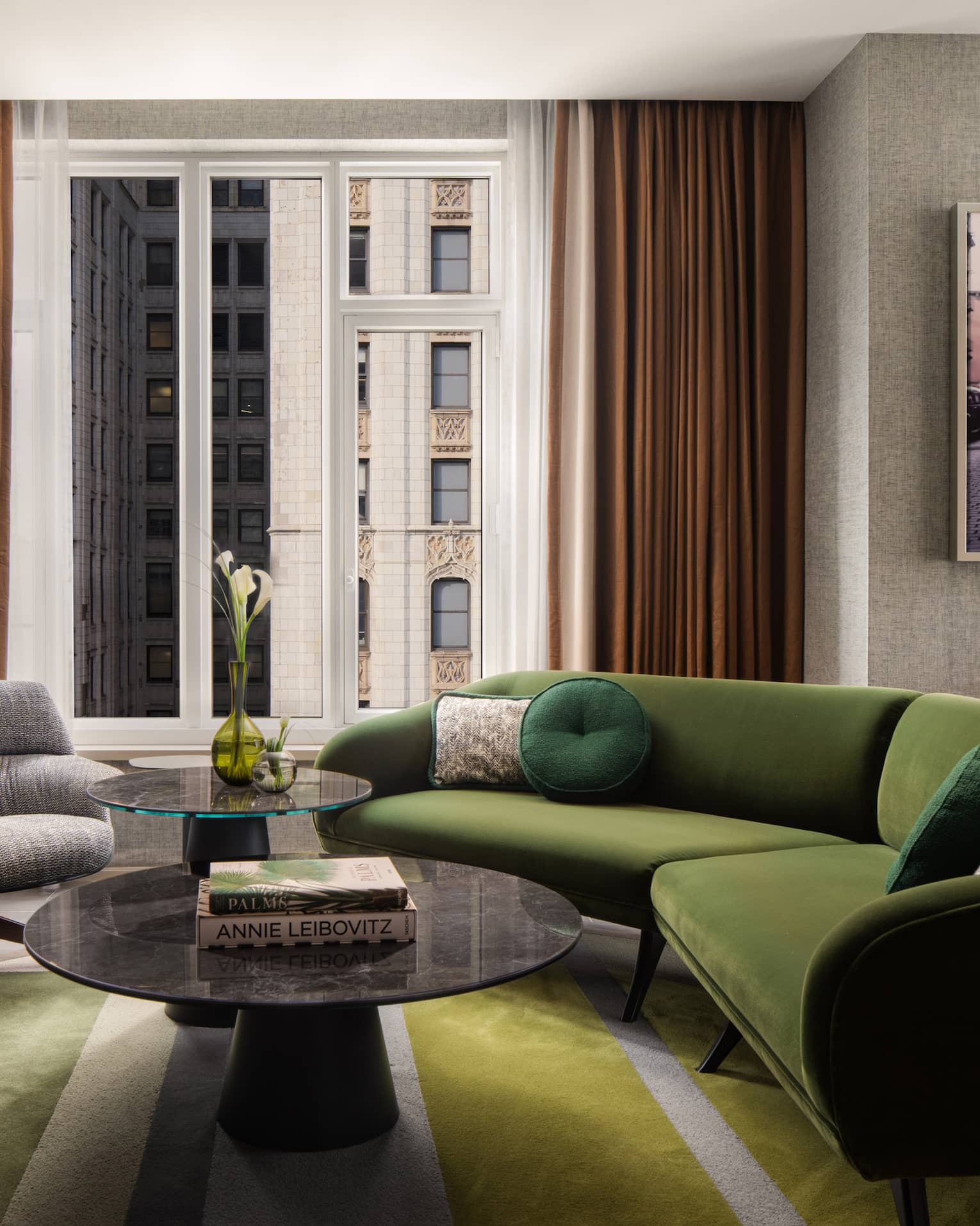 Suite living room with green sectional sofa, round coffee tables, gray armchair, wall art, and large window overlooking city buildings