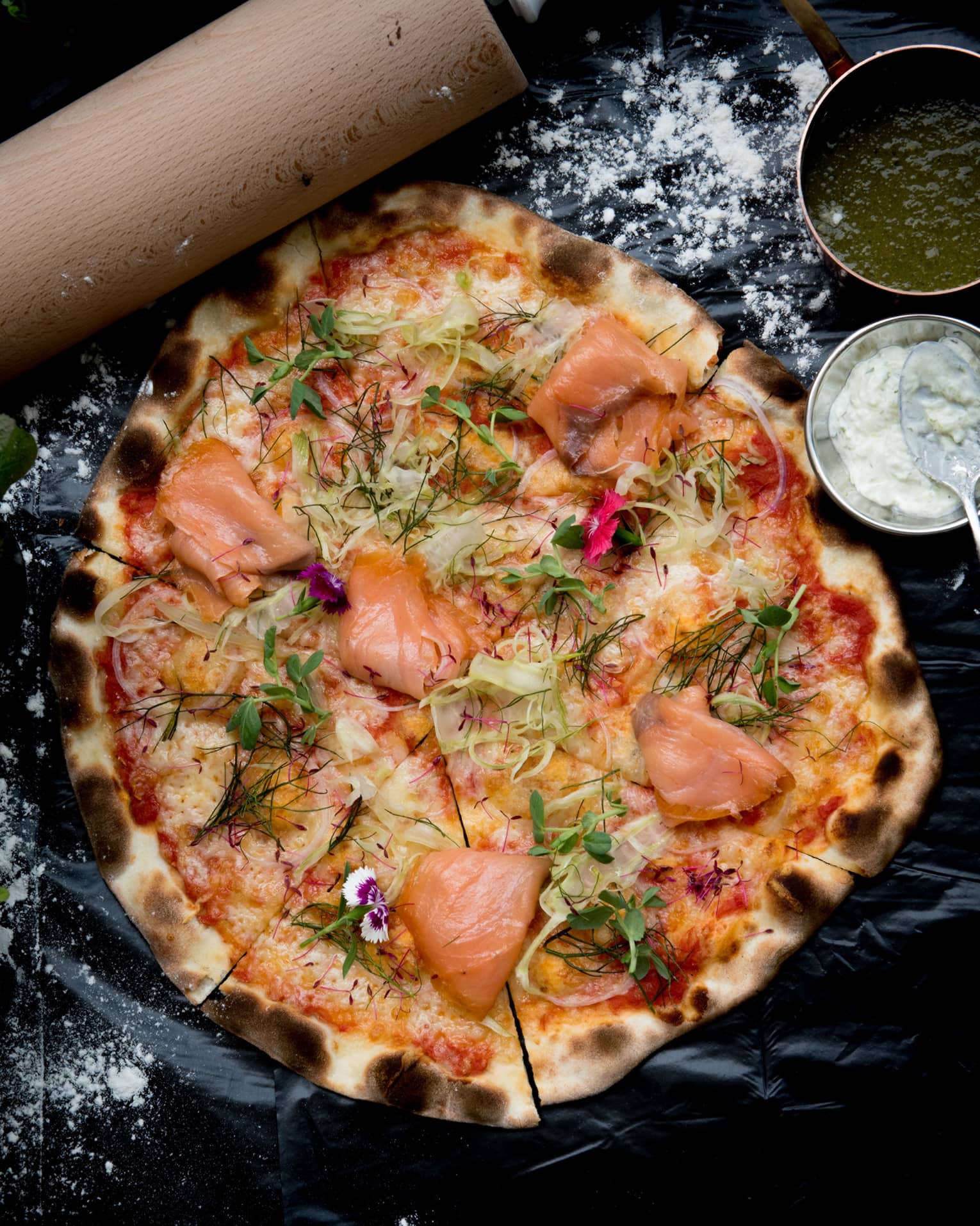 A wood-fired pizza topped with smoked salmon, fennel and dill, bowls of green and white sauces on the side.