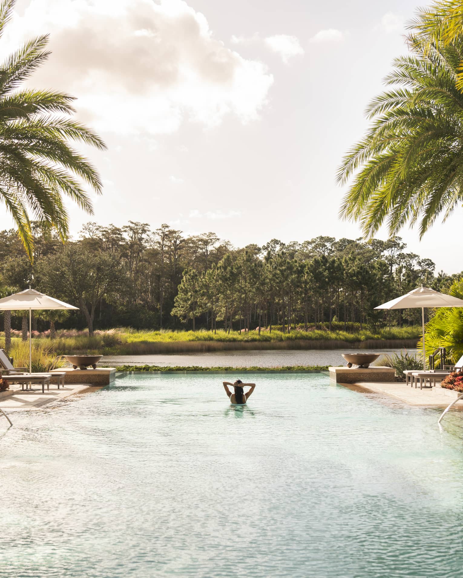 A woman alone in a large pool with lounge chairs and palm trees around it.
