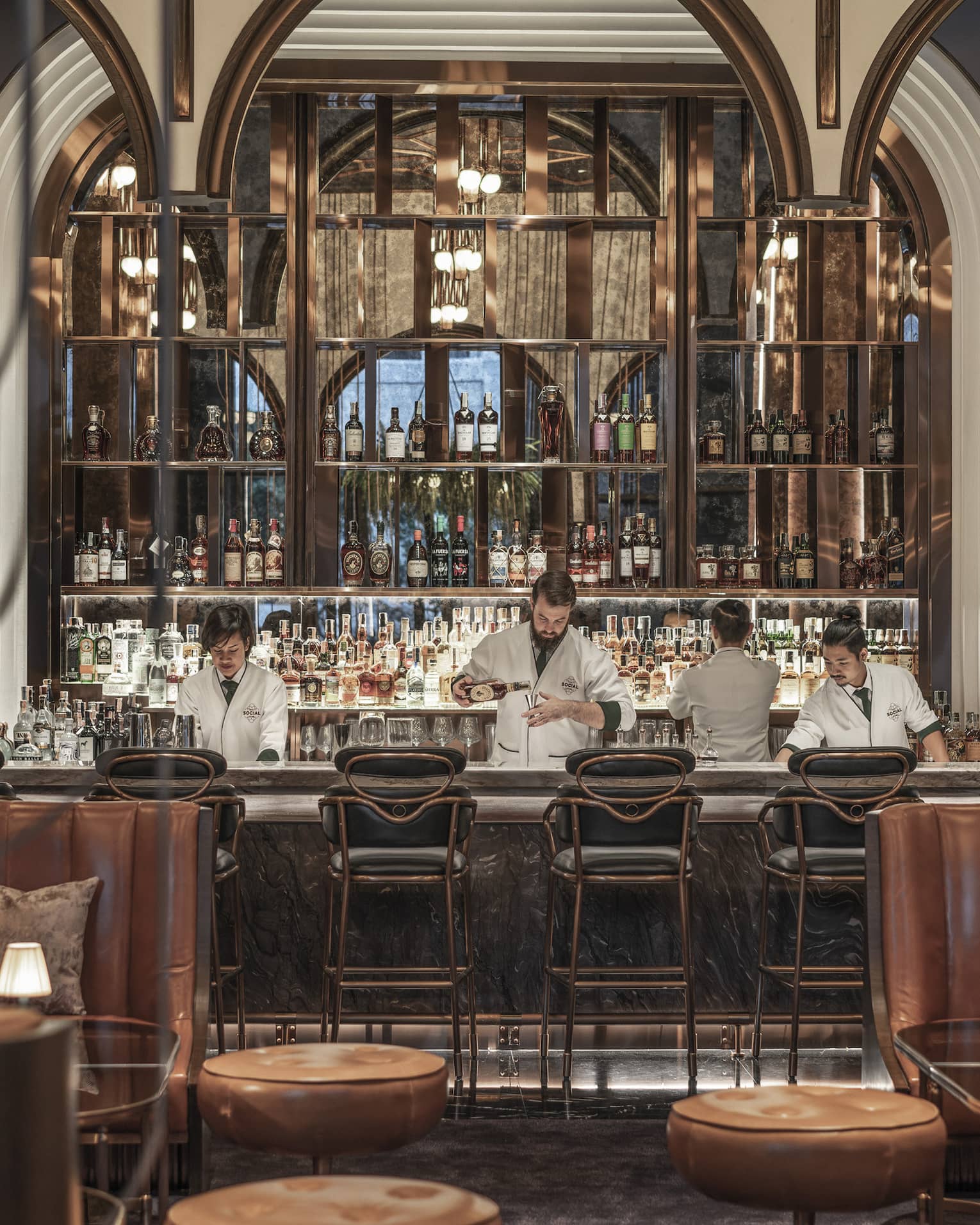 Chic bar with leather seating, plants on the walls and a well-stocked backlit bar, where bartenders in white jackets prepare drinks behind the counter.