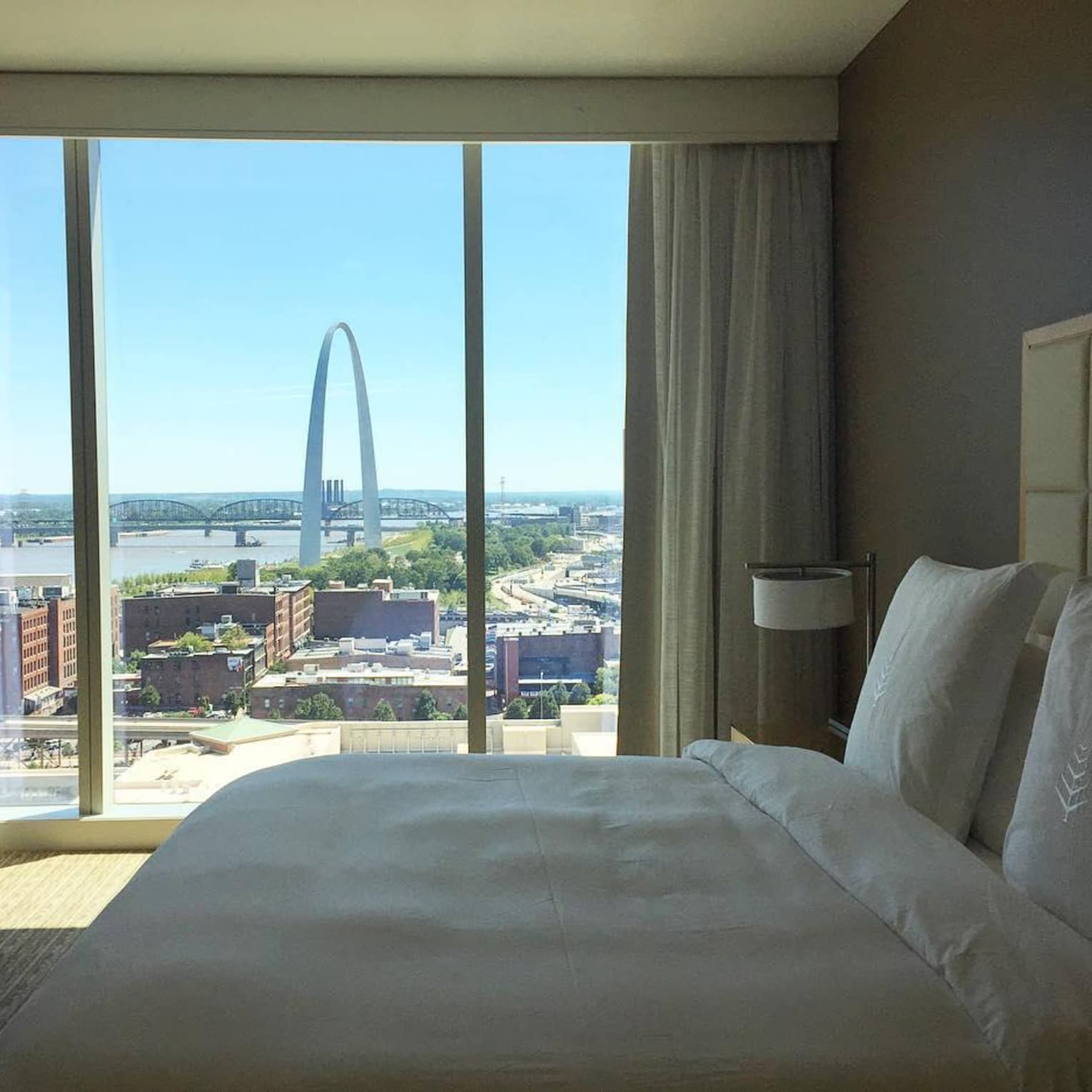 Luxury hotel room with a large window view of the St. Louis Gateway Arch, featuring a neatly made bed with white linens and natural light streaming in