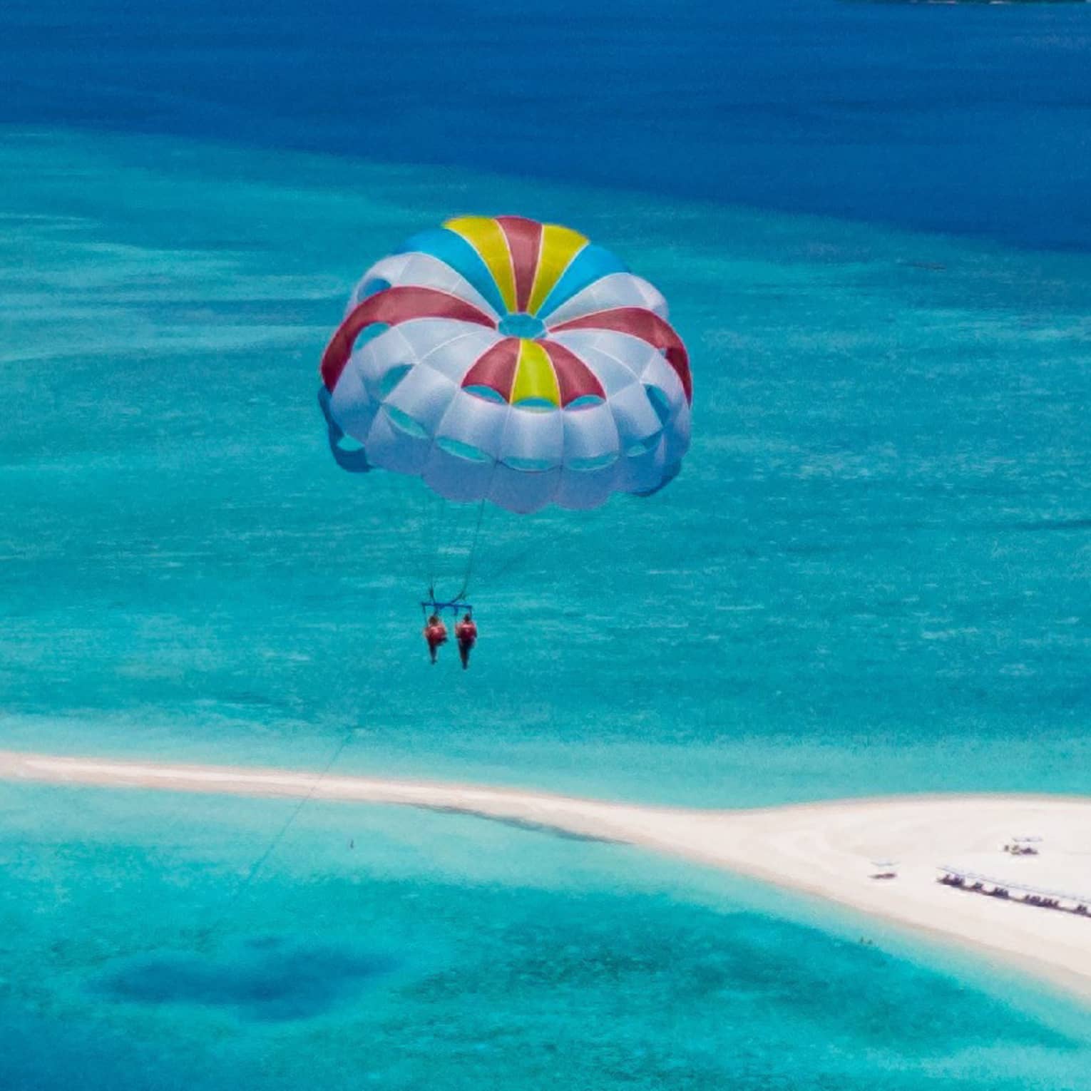 Aerial view of two people parasailing in tandem, colourful sail floating above an expanse of aqua water and a white sandbar.