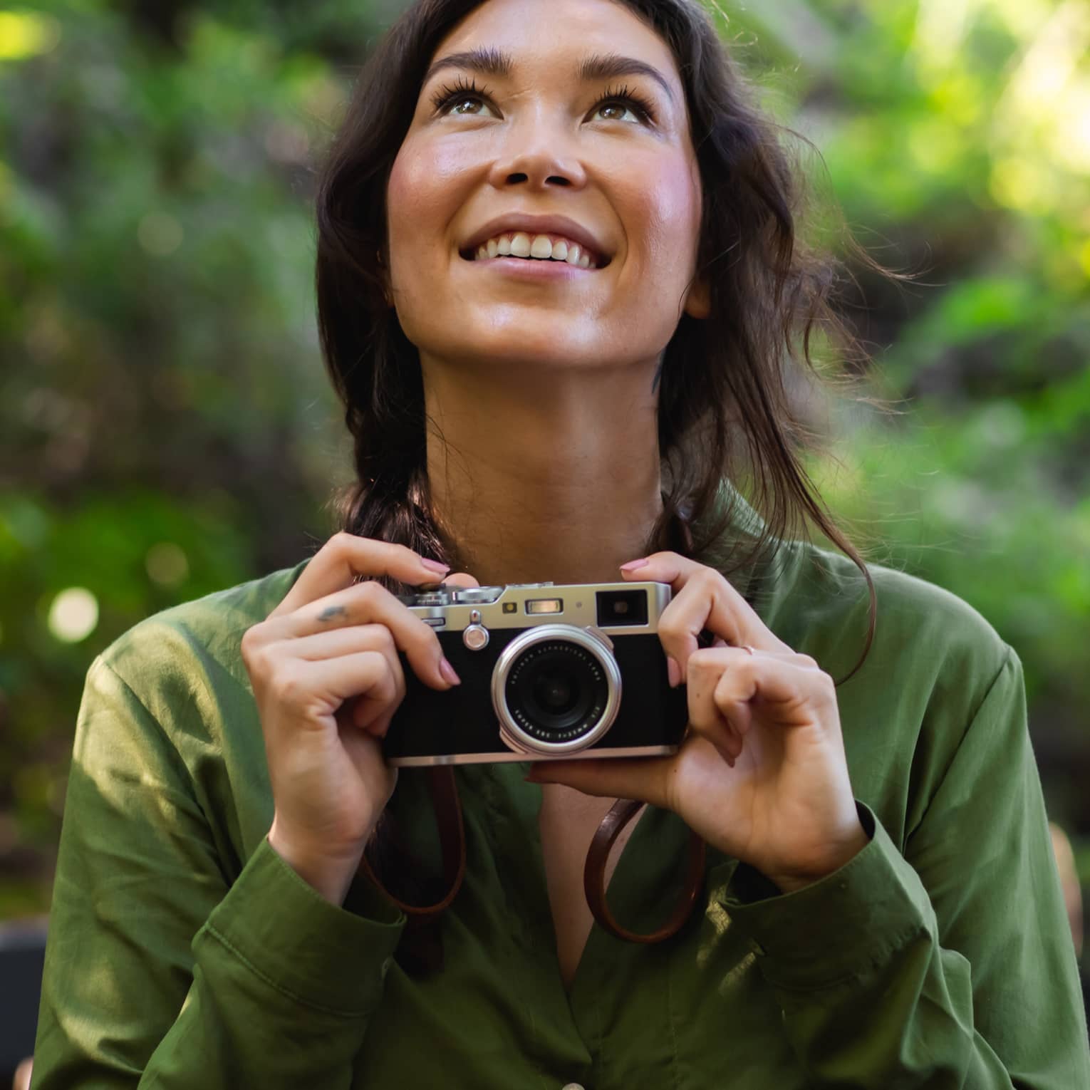 Woman with long dark hair wearing a green button-down shirt holds a camera as she looks upward in the forest