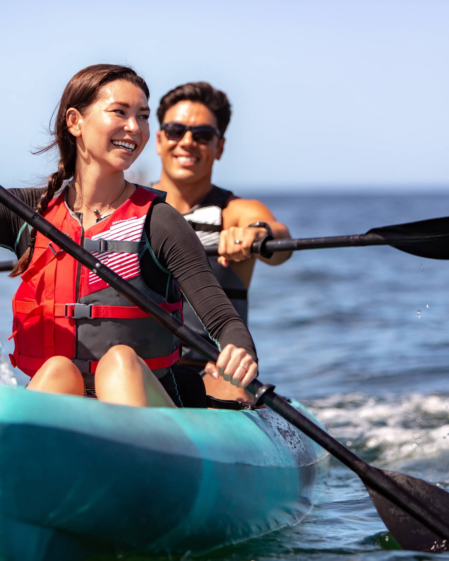 Close-up front view of two smiling adults paddling in a tandem kayak on open water under a sunny blue sky.