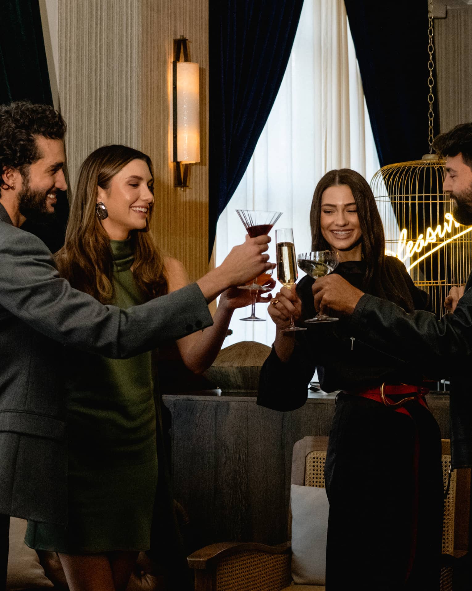 Four people raise their glasses in a toast in a cozy, softly lit lounge setting.