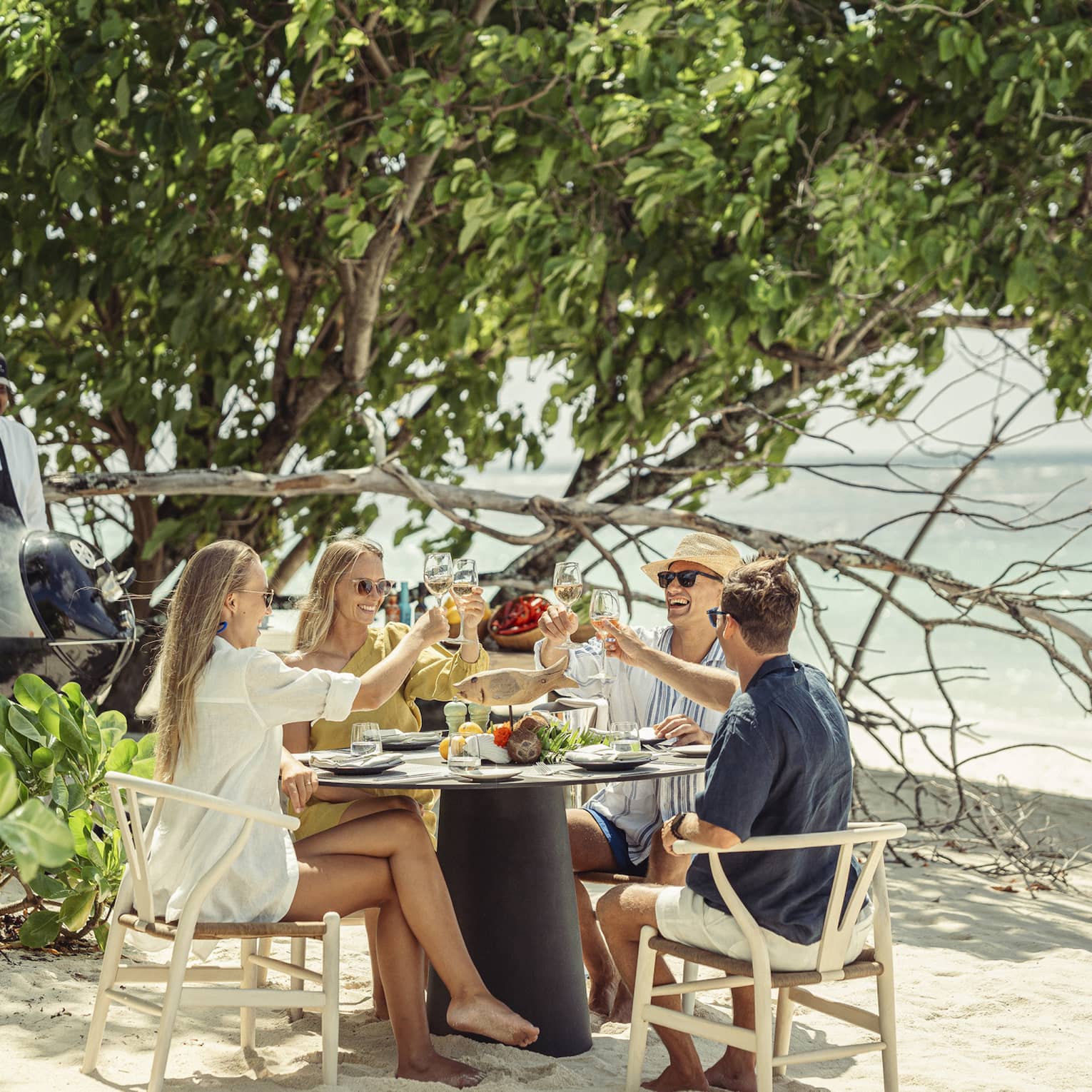 A group sits at a round table for a beach-side meal, clinking glasses while a smiling chef cooks on a coal barbecue nearby.