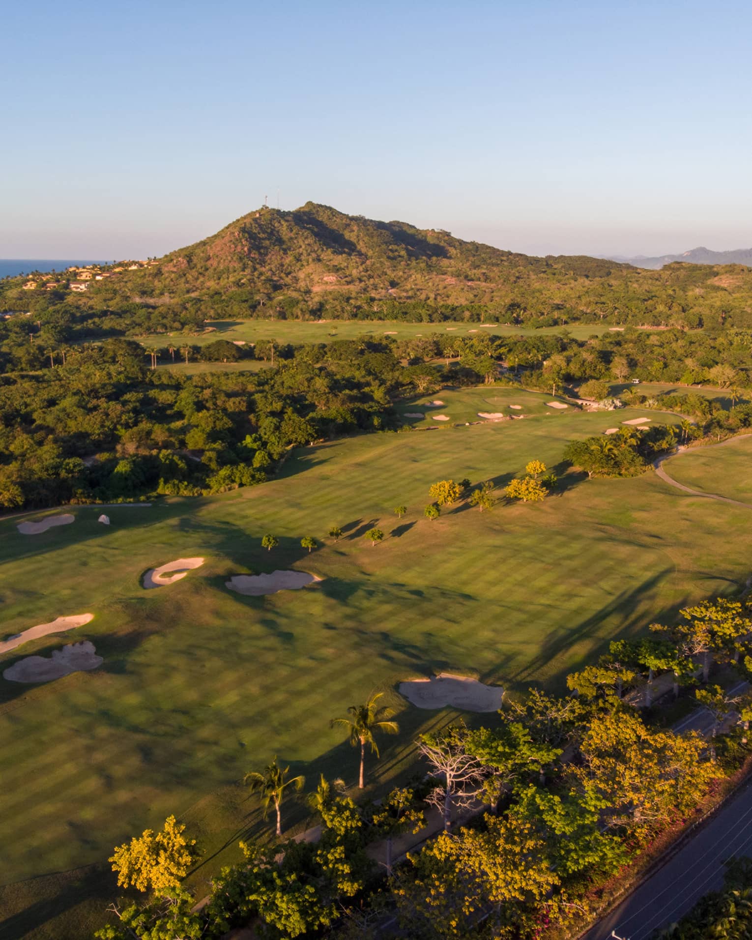 Aerial view of a golf course over the rolling coastal landscape; houses rising along the hillside, ocean in the distance.