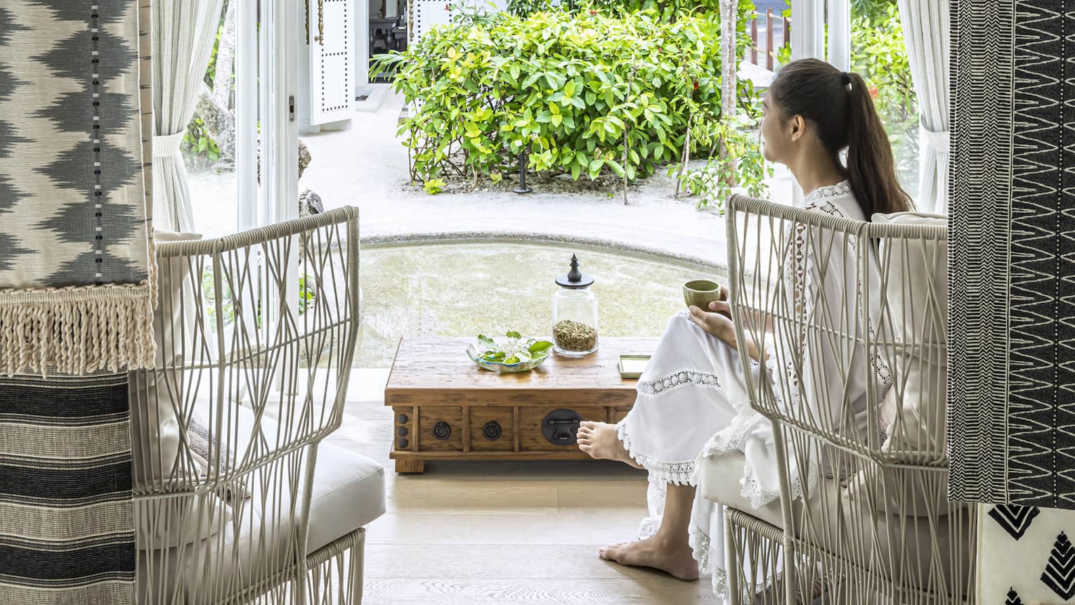 A woman sits in white wicker chair in the four seasons maldives spa waiting area and overlooks the garden outside an opened window