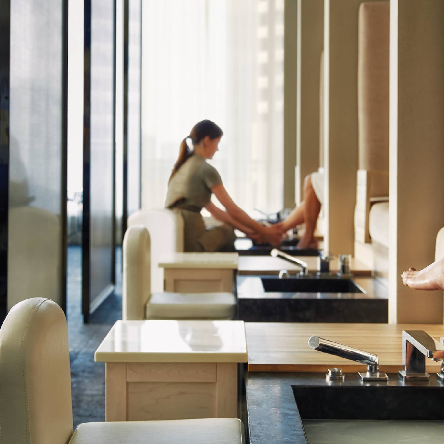 Woman's feet on pedicure sink along row of spa chairs, salon staff in background