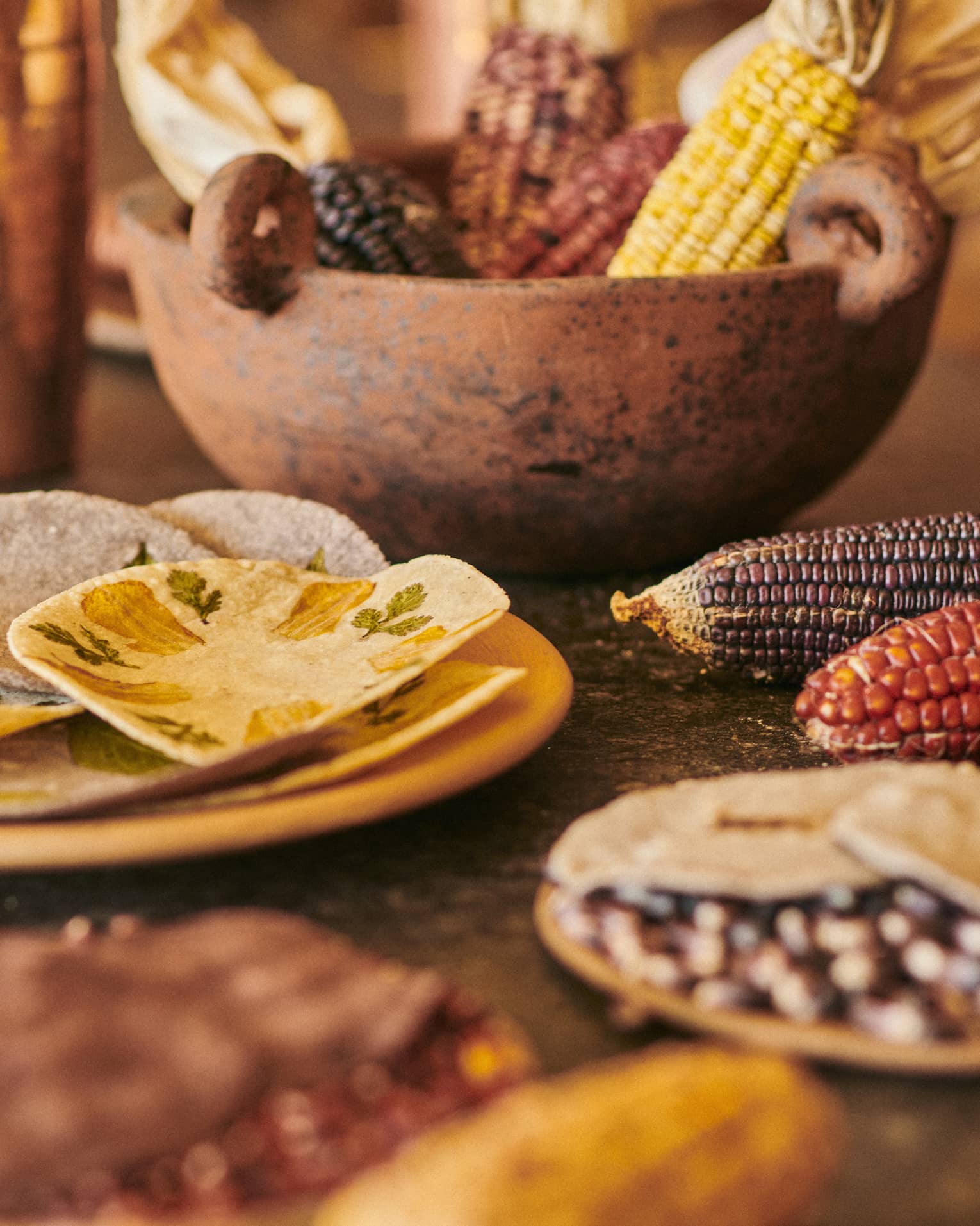 Close-up of copper cups, a rust-coloured stone bowl holding dried corn cobs and a yellow plate with hand-pressed tortillas.
