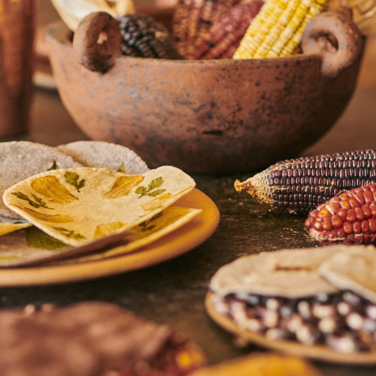 Close-up of copper cups, a rust-coloured stone bowl holding dried corn cobs and a yellow plate with hand-pressed tortillas.