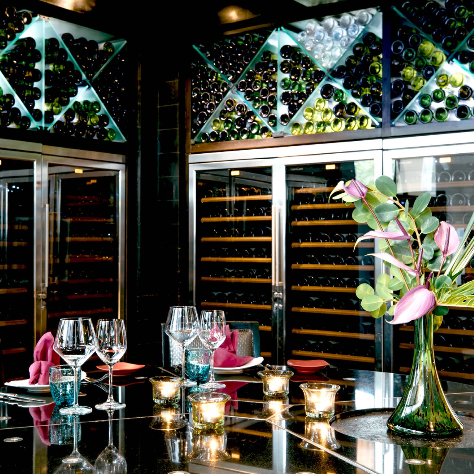 Opulent setting of goblets, candles and a tropical flower atop a mirrored table, surrounded by floor-to-ceiling wine racks.