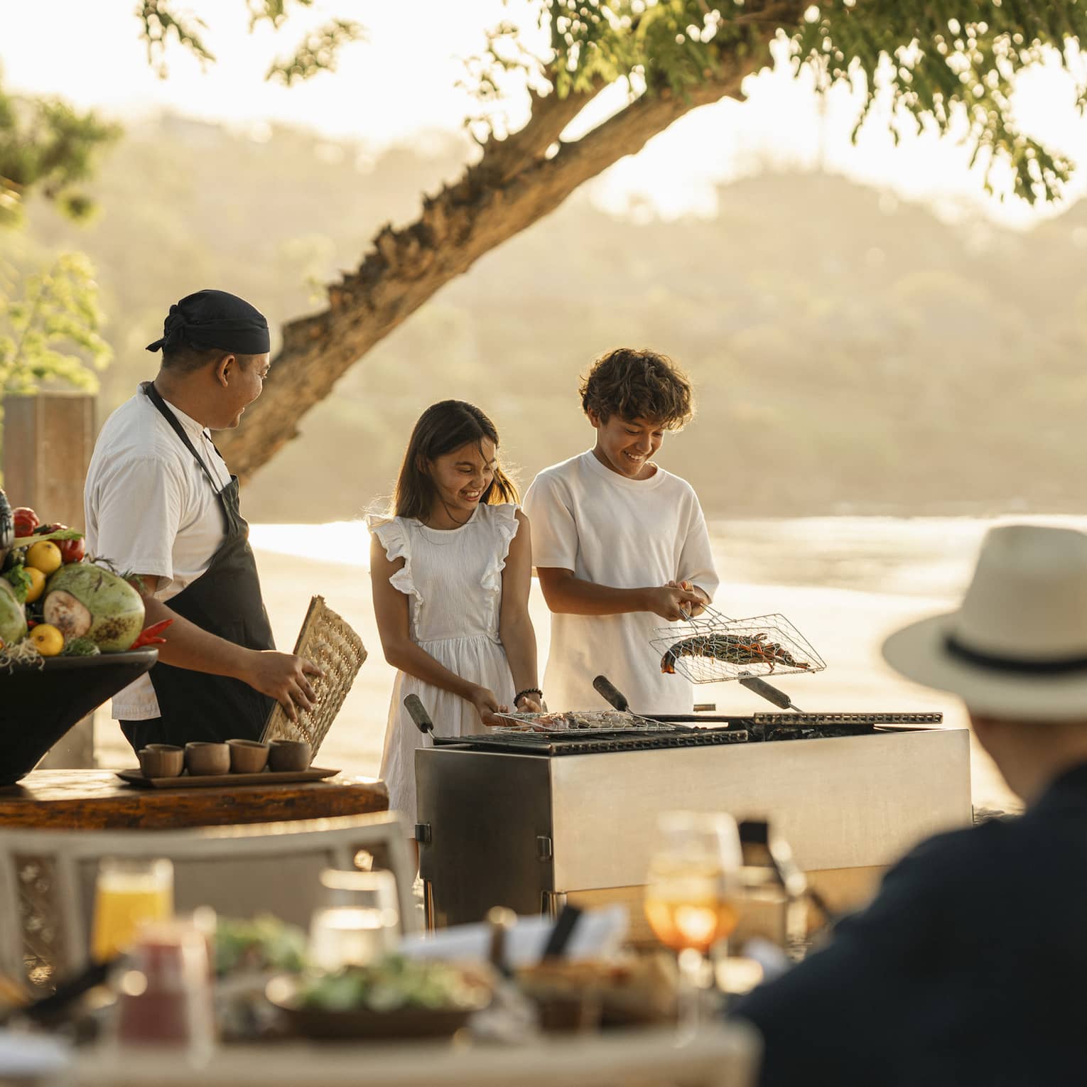 A chef smiles at two children who barbecue shellfish in a grilling basket on the beach as their parents watch.