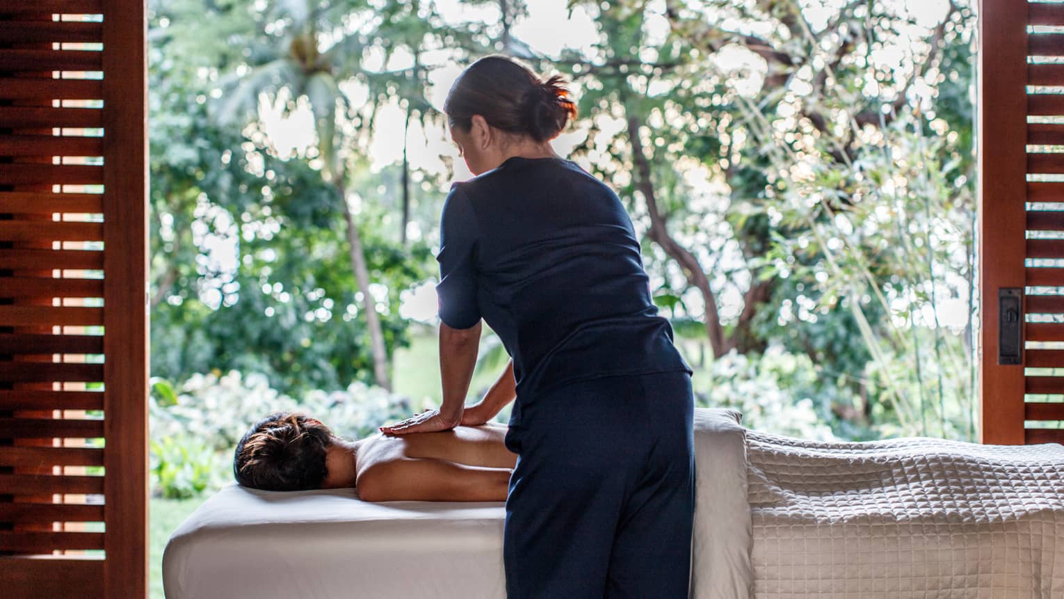 Spa staff massages woman's bare shoulders as she lies under sheet on massage table