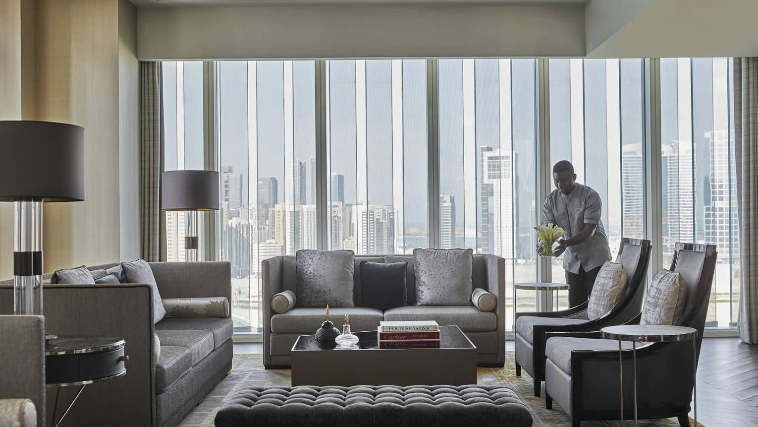 Spacious hotel living room with gray sofas, armchairs, a tufted ottoman, floor-to-ceiling windows offering city views and a staff member arranging flowers