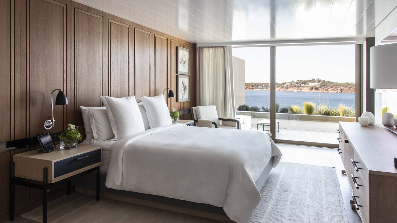 White king bed, wooden accent wall, contemporary night stand and dresser, wall of windows with sea view