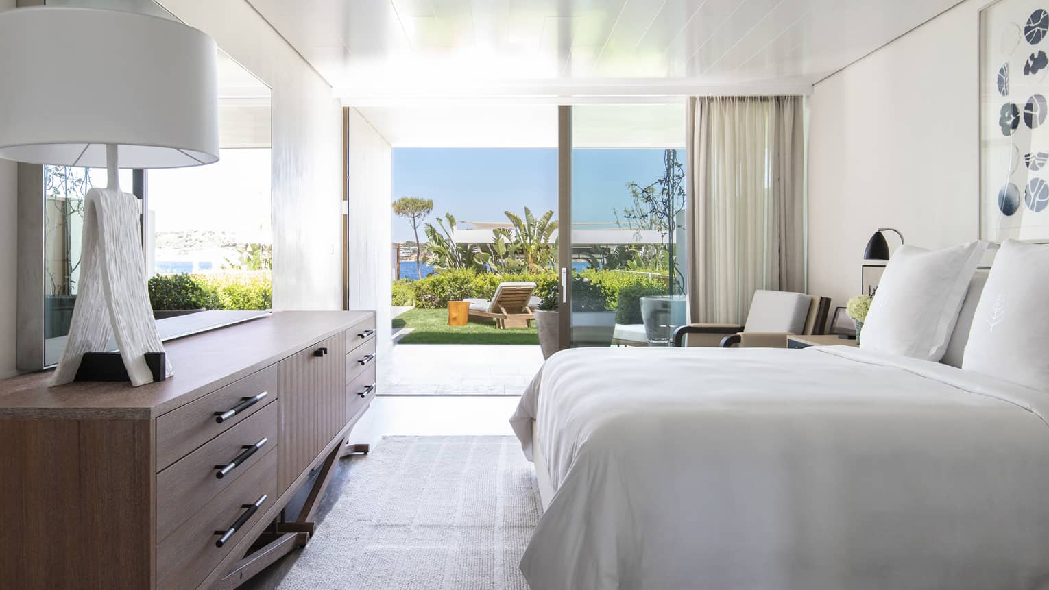 Bedroom with plush king bed, modern wooden dresser with lamp, walk-out terrace to garden