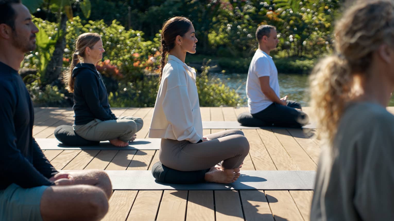 A group of people sit cross legged on wooden deck in garden setting, practicing yoga