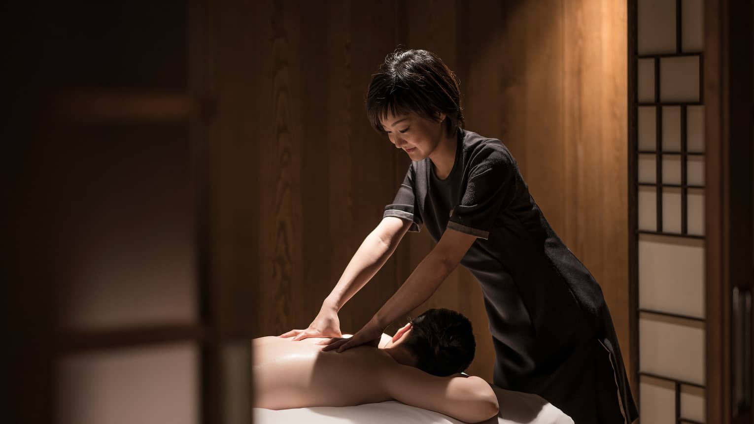 Man lies face-down on massage table as masseuse in black uniform rubs his shoulders
