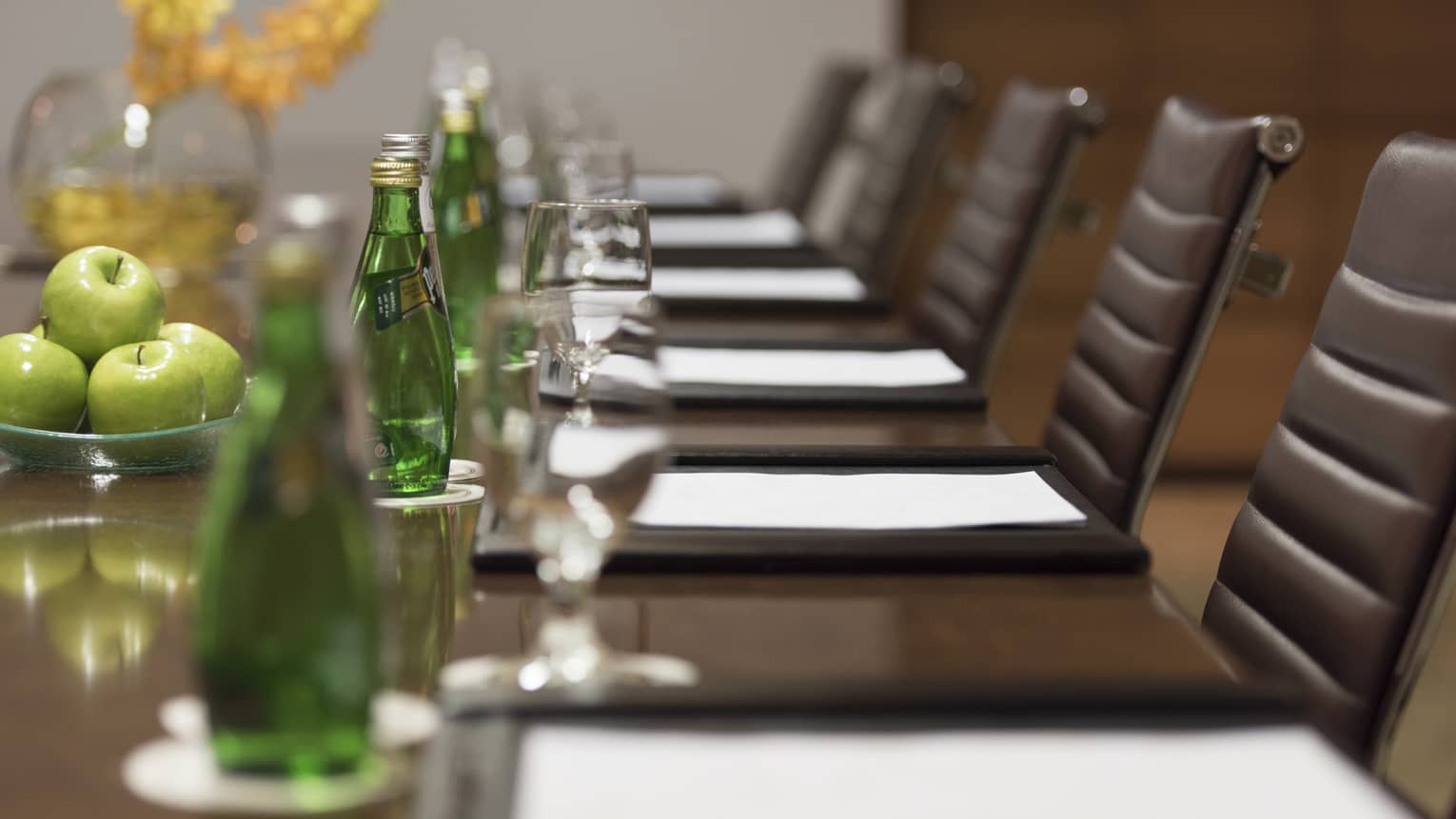 Close-up of business meeting table row of chairs, paper agendas, green glass bottles, apples