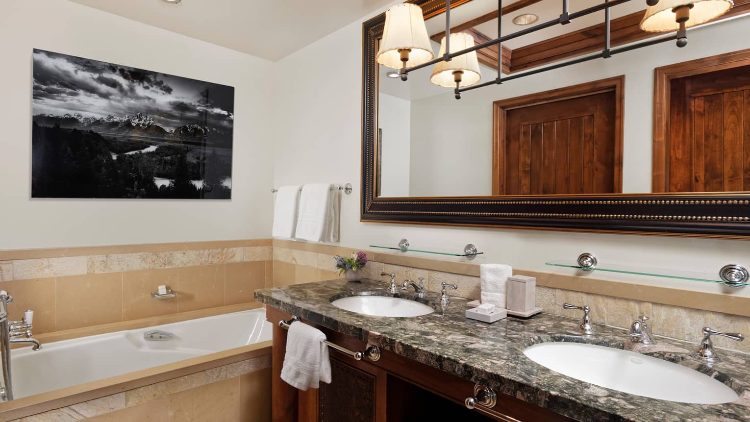 Luxury resort bathroom with granite double vanity, large framed mirror, beige tiled bathtub and a black-and-white landscape photograph on the wall