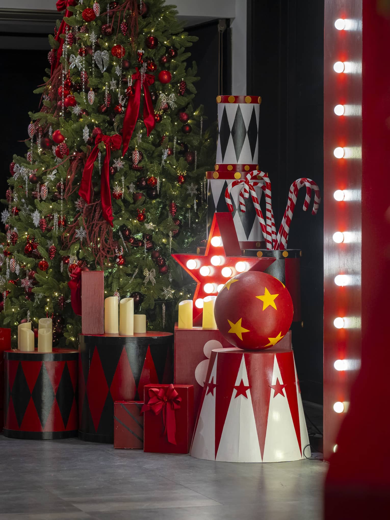 In a dark room, a tall Christmas tree is surrounded by candles, large candy canes, and wooden boxes, cylinders and balls.