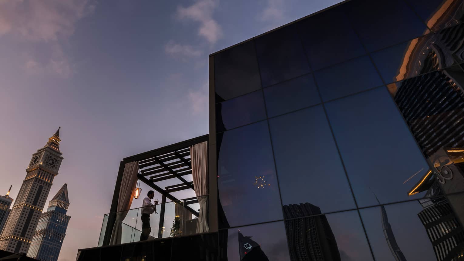Silhouette of man with drink in glass corner balcony of Luna Sky Bar at dusk, Dubai towers
