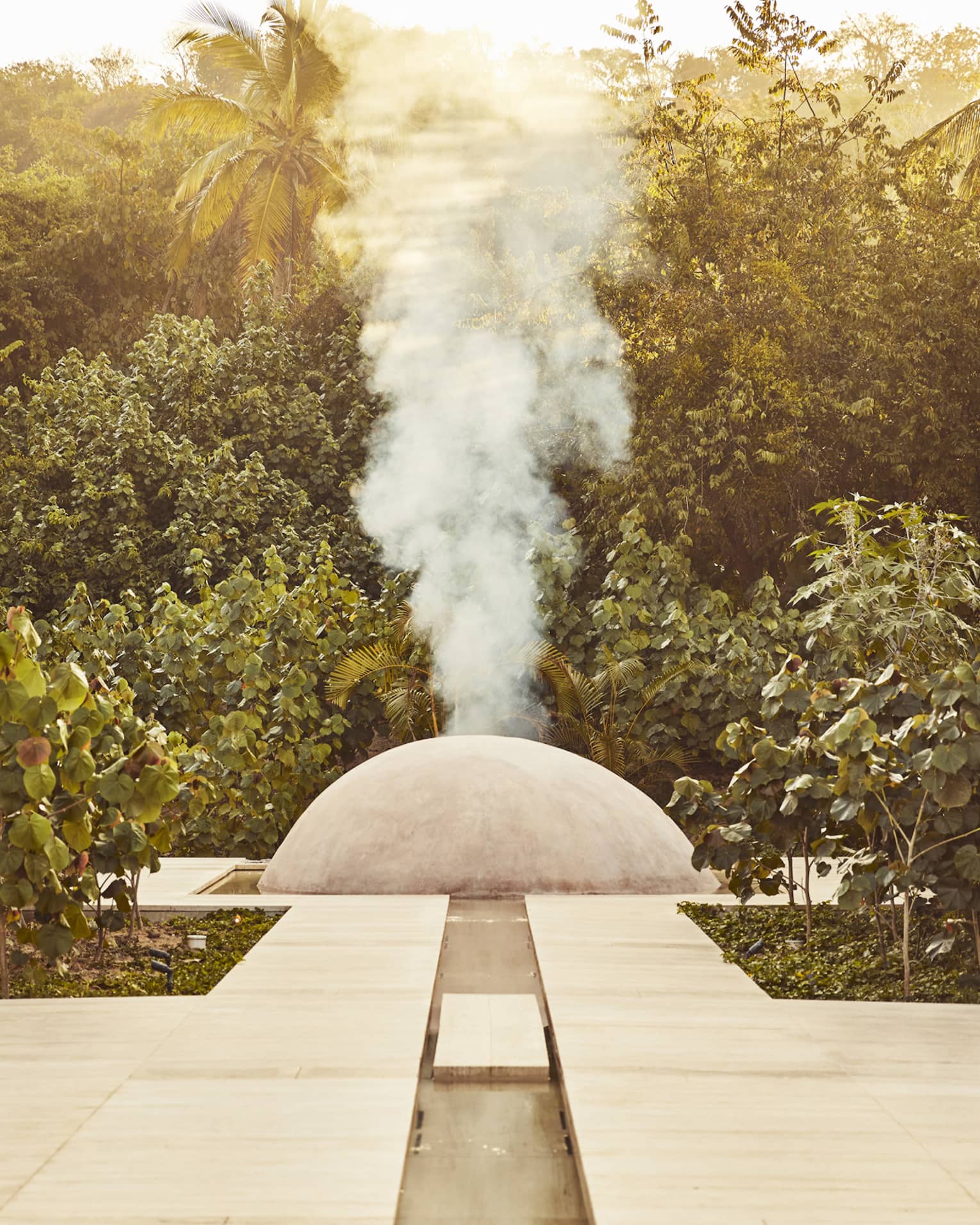 White steam rises from a small cement dome nestled between two large stone walls and surrounded by dense dark-green foliage.