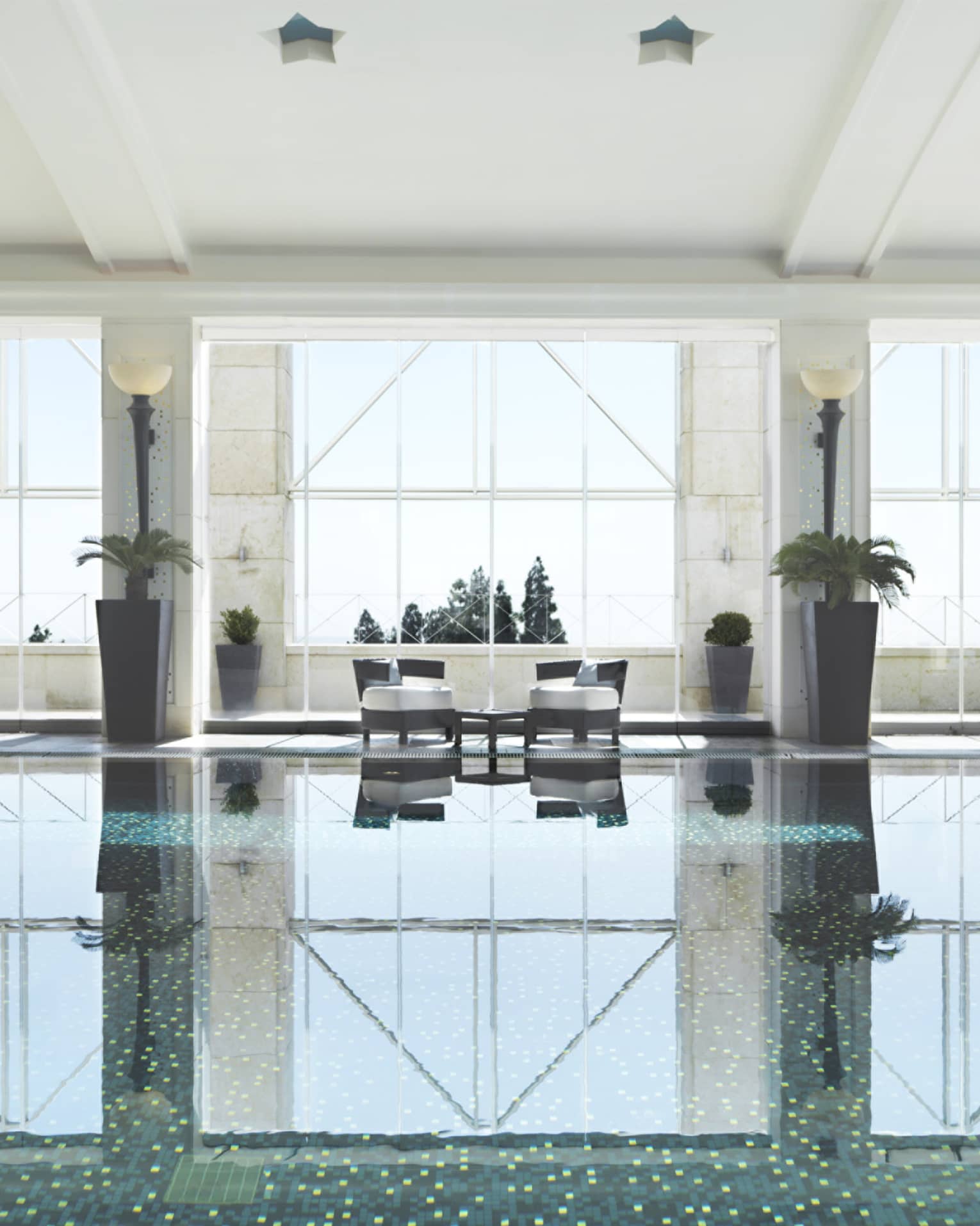 Indoor swimming pool reflecting light of three tall windows, white walls and pool chairs