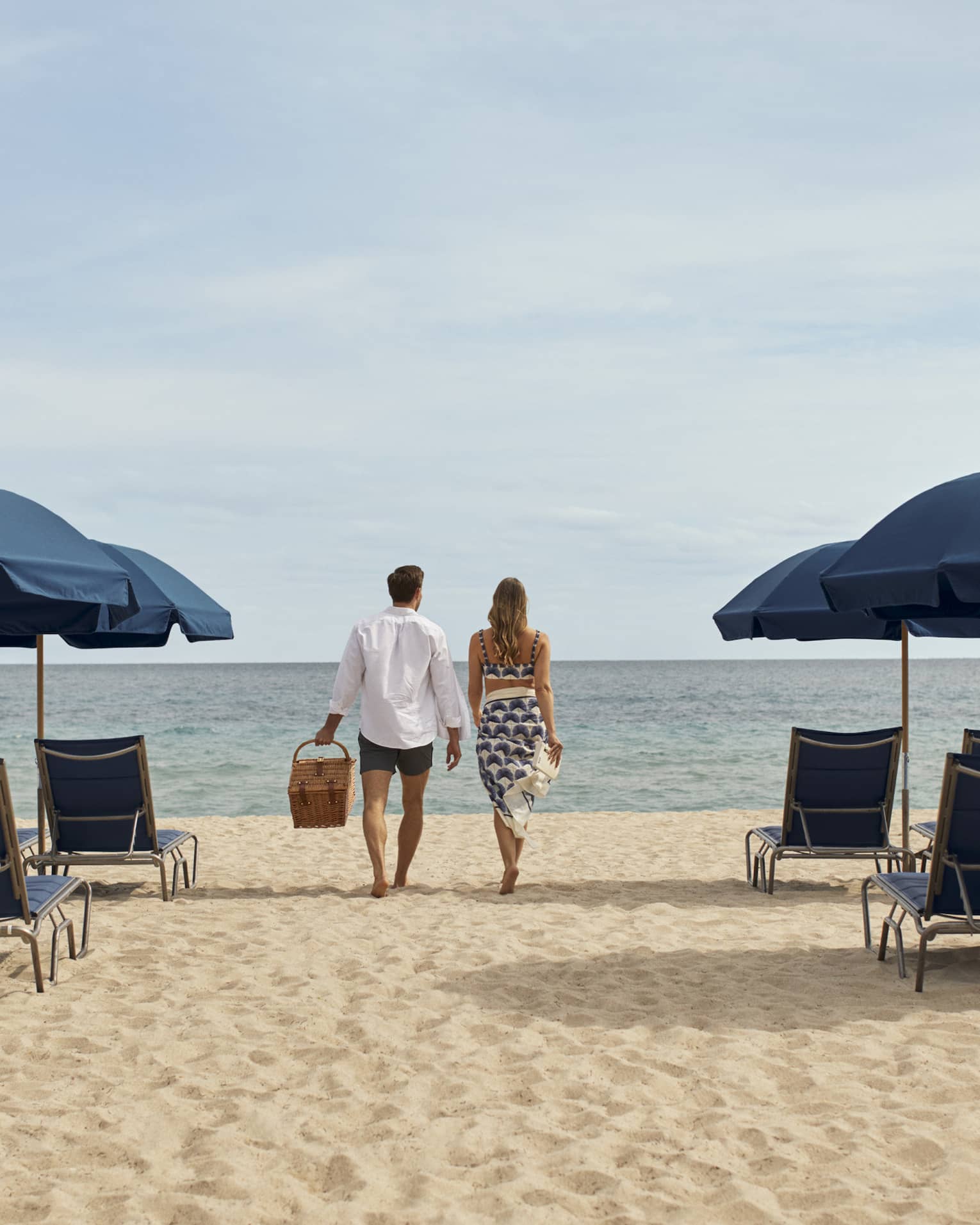 A man and woman walking between blue lounge chairs and umbrellas on sand towards the ocean.