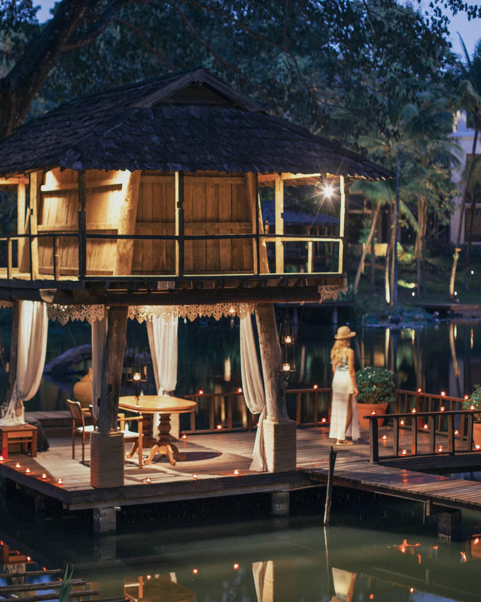 Person standing on a dock beside a candlelit lakeside pavilion with a thatched roof and draped curtains at dusk.