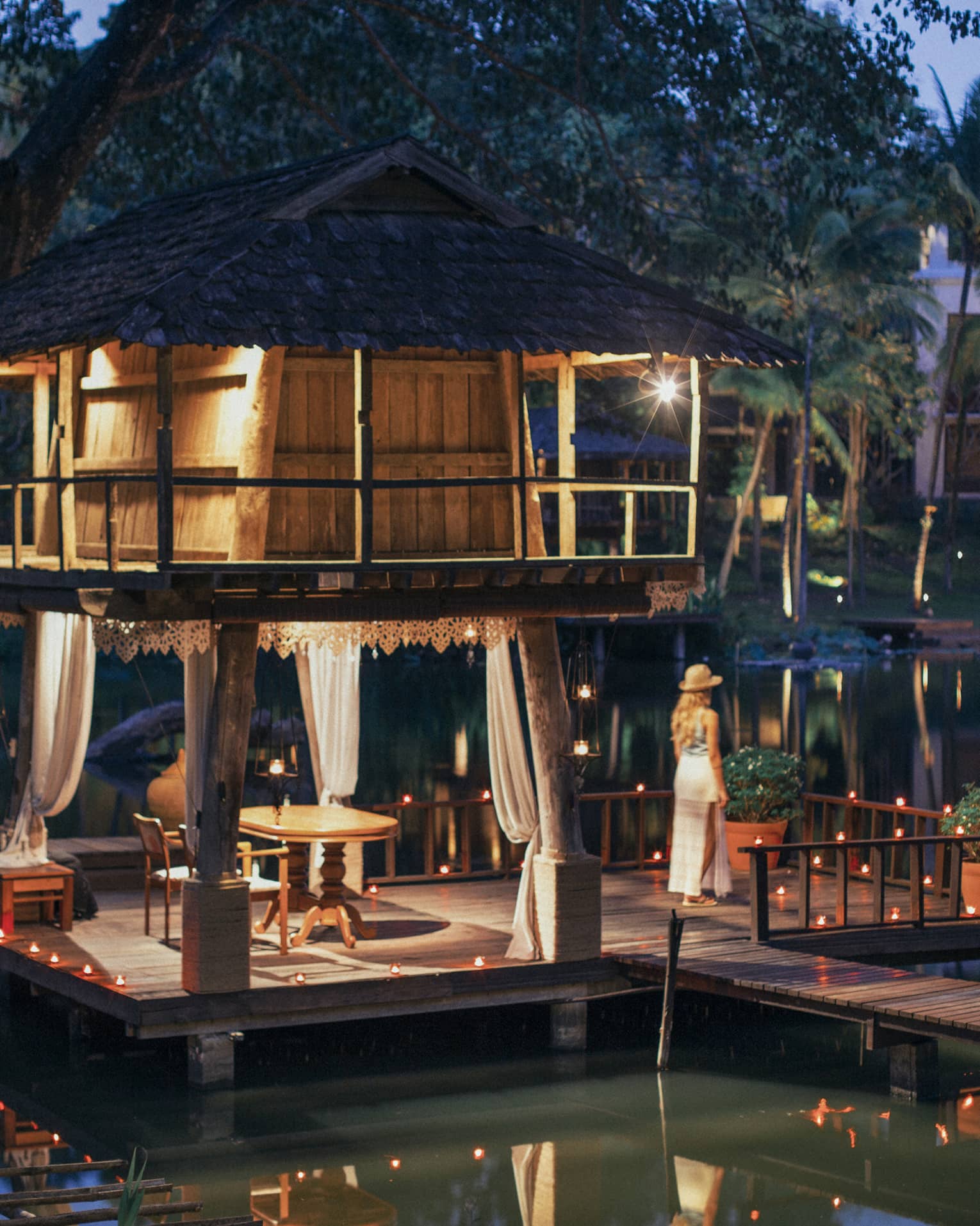 Person standing on a dock beside a candlelit lakeside pavilion with a thatched roof and draped curtains at dusk.