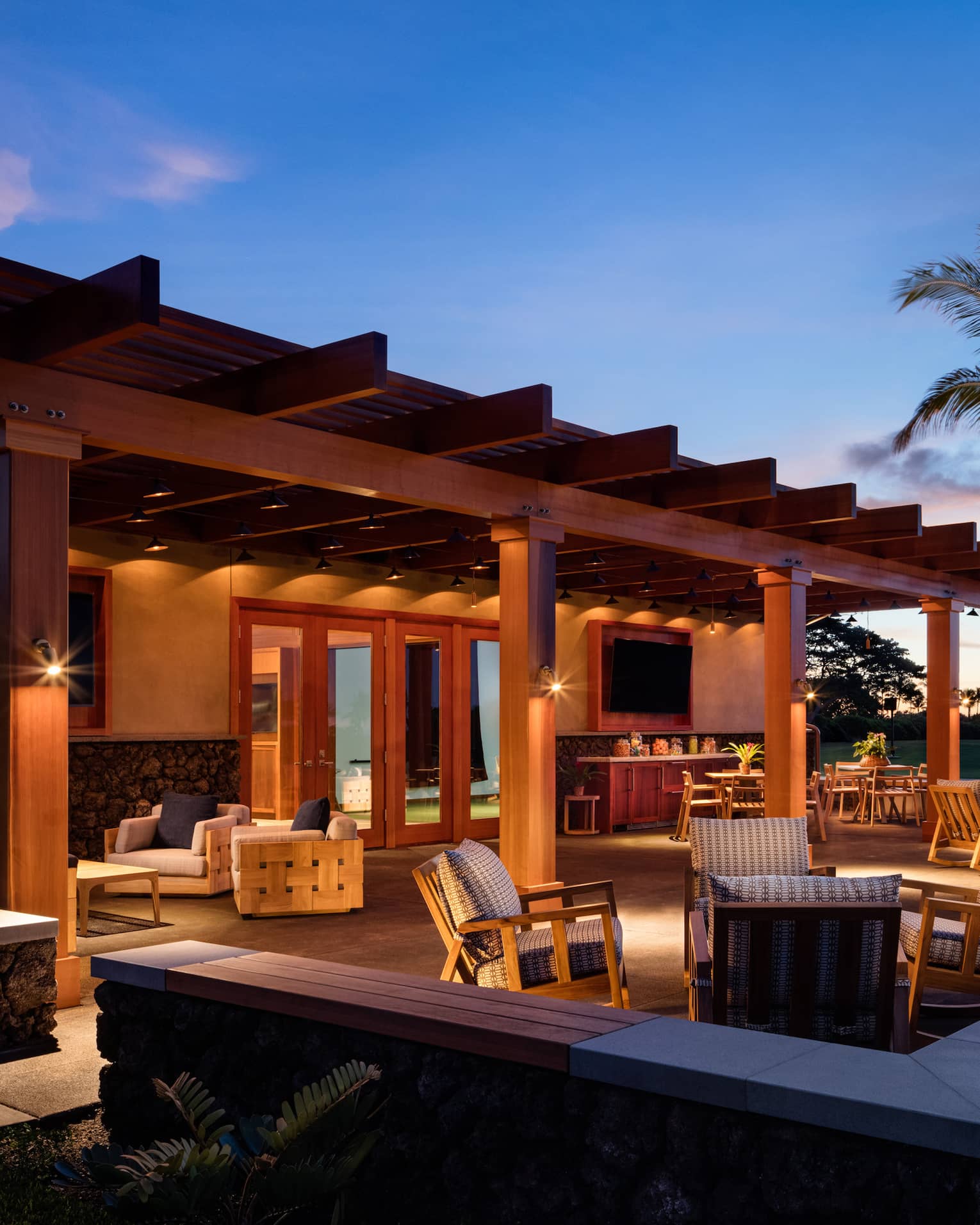 A warmly lit patio furnished with tables and cushioned armchairs under a clear, royal-blue night sky, sunset in the distance.