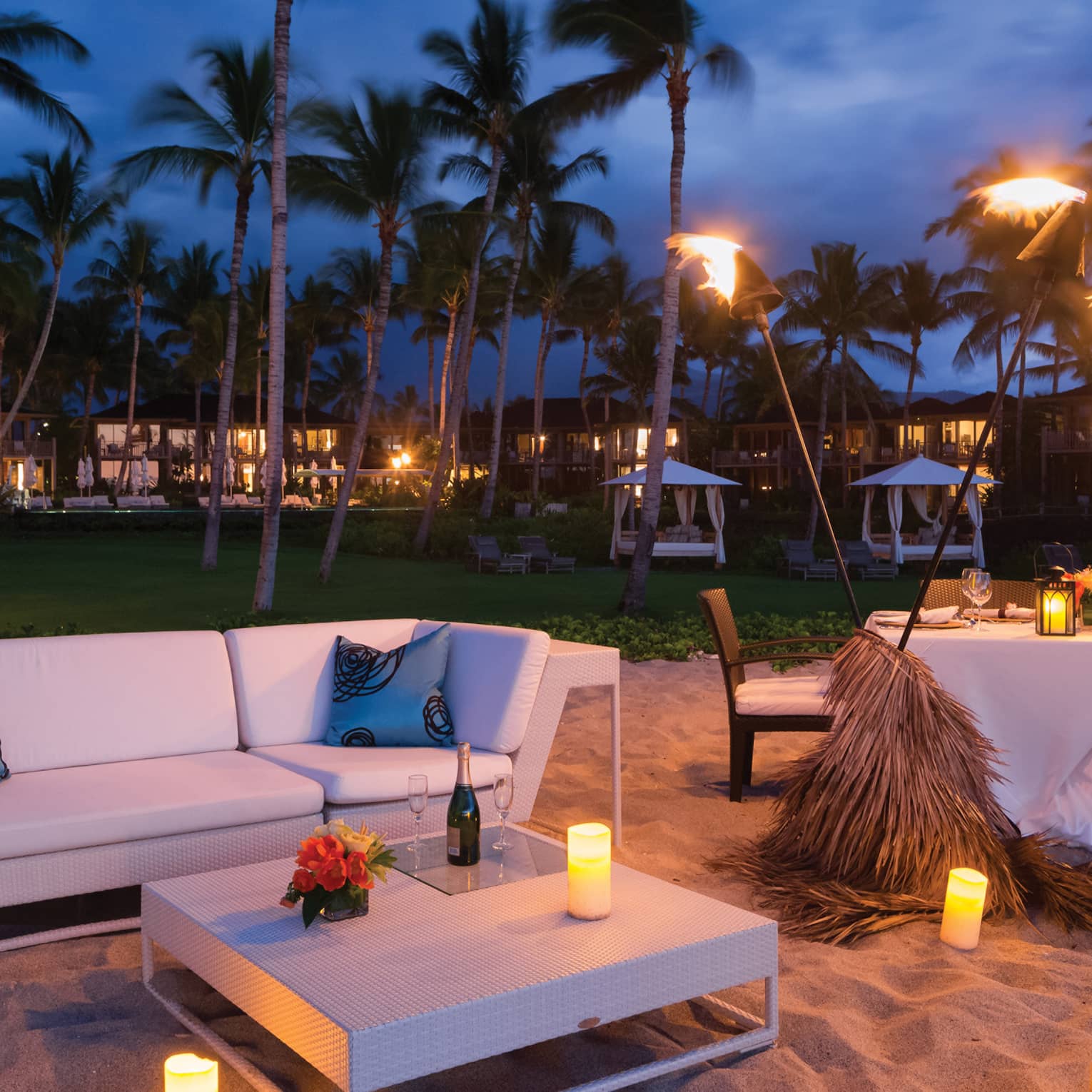 A sofa, coffee table, and dinner for two on a beach lit by the glow of torches and candles, palm trees swaying in the wind.