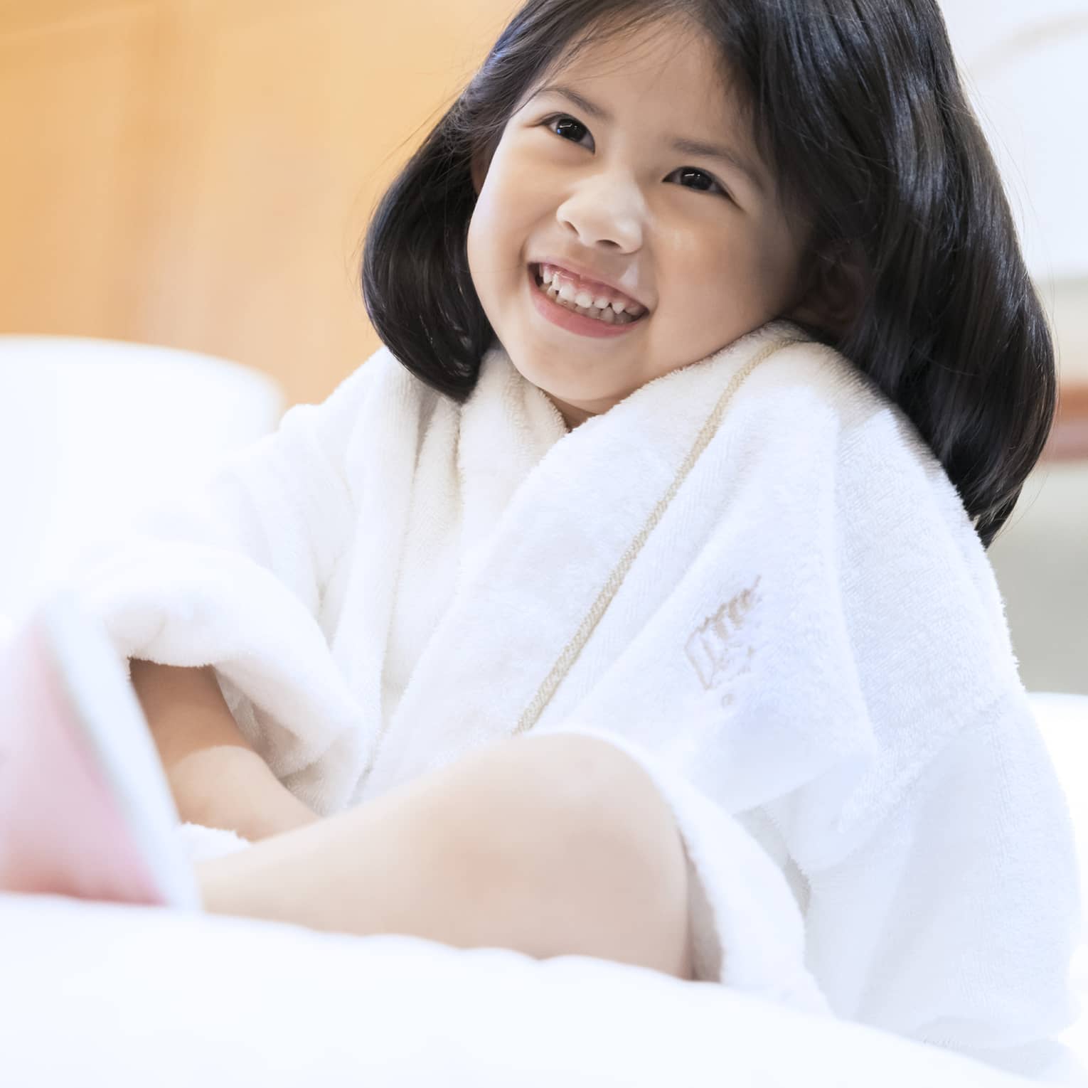 Smiling little girl wearing white bath robe and pink slippers sits on bed
