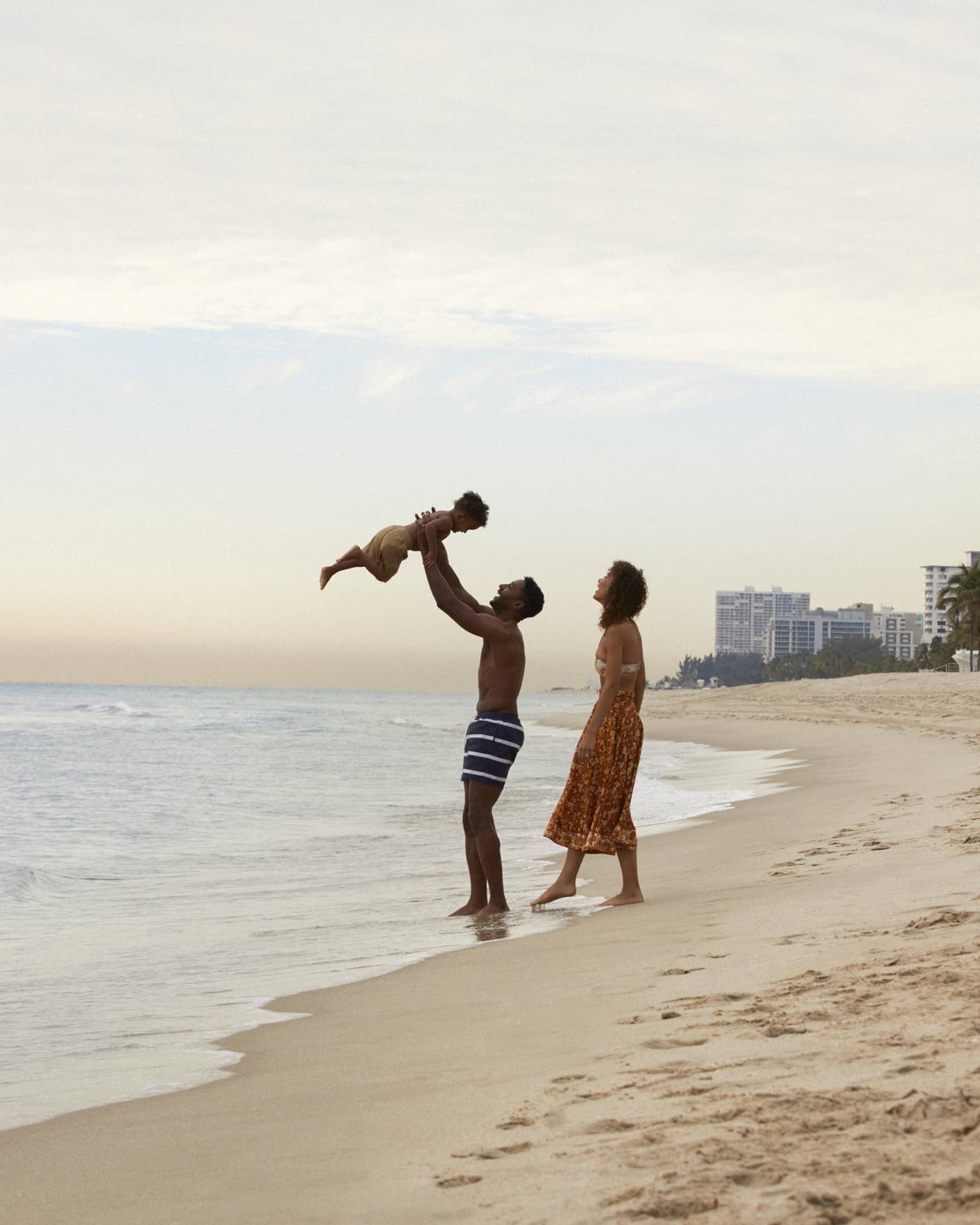 A mother and father and son at the beach, the father is holding the sun up in the air.
