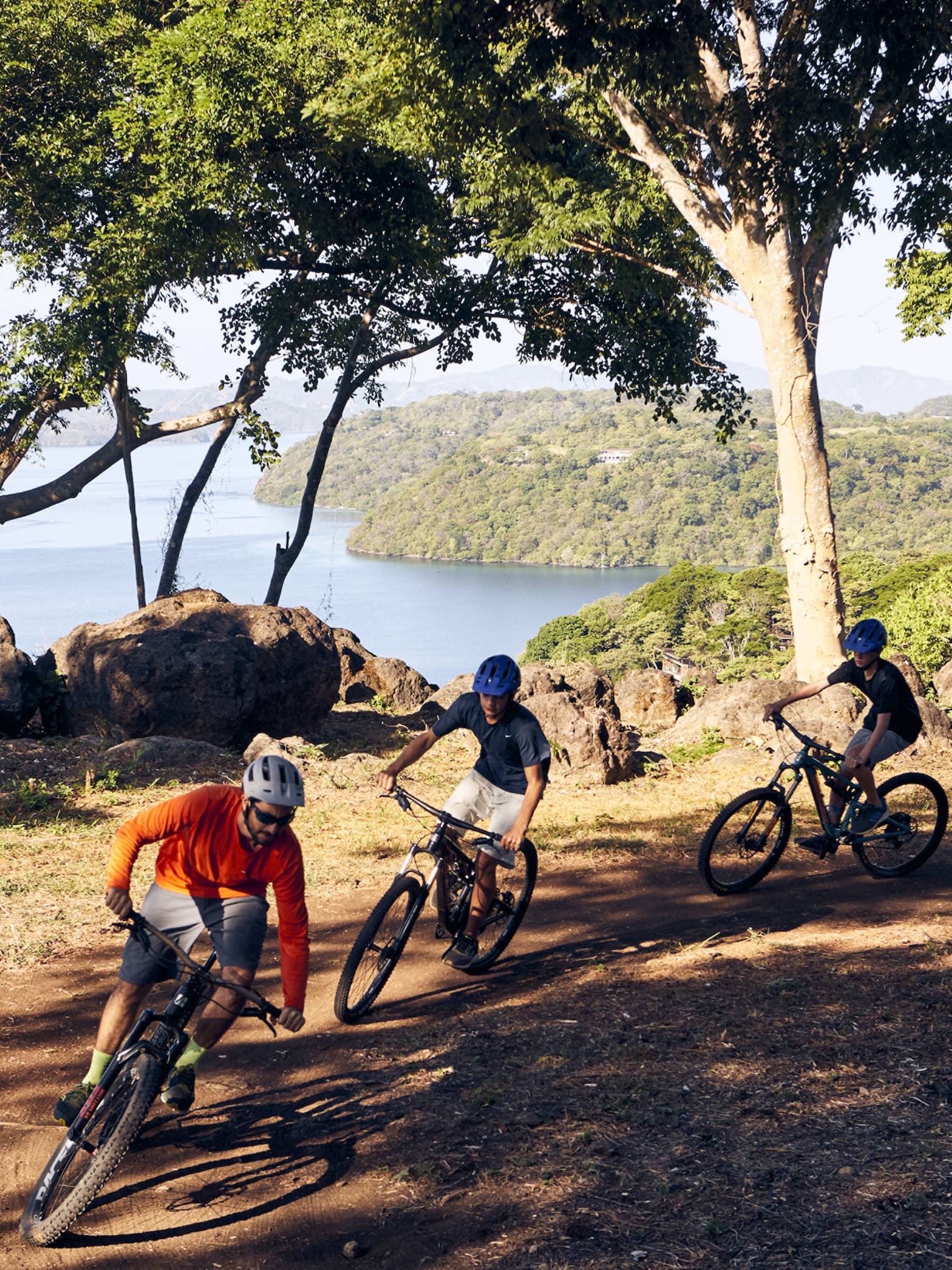 Three people ride mountain bikes along a hillside path overlooking the ocean
