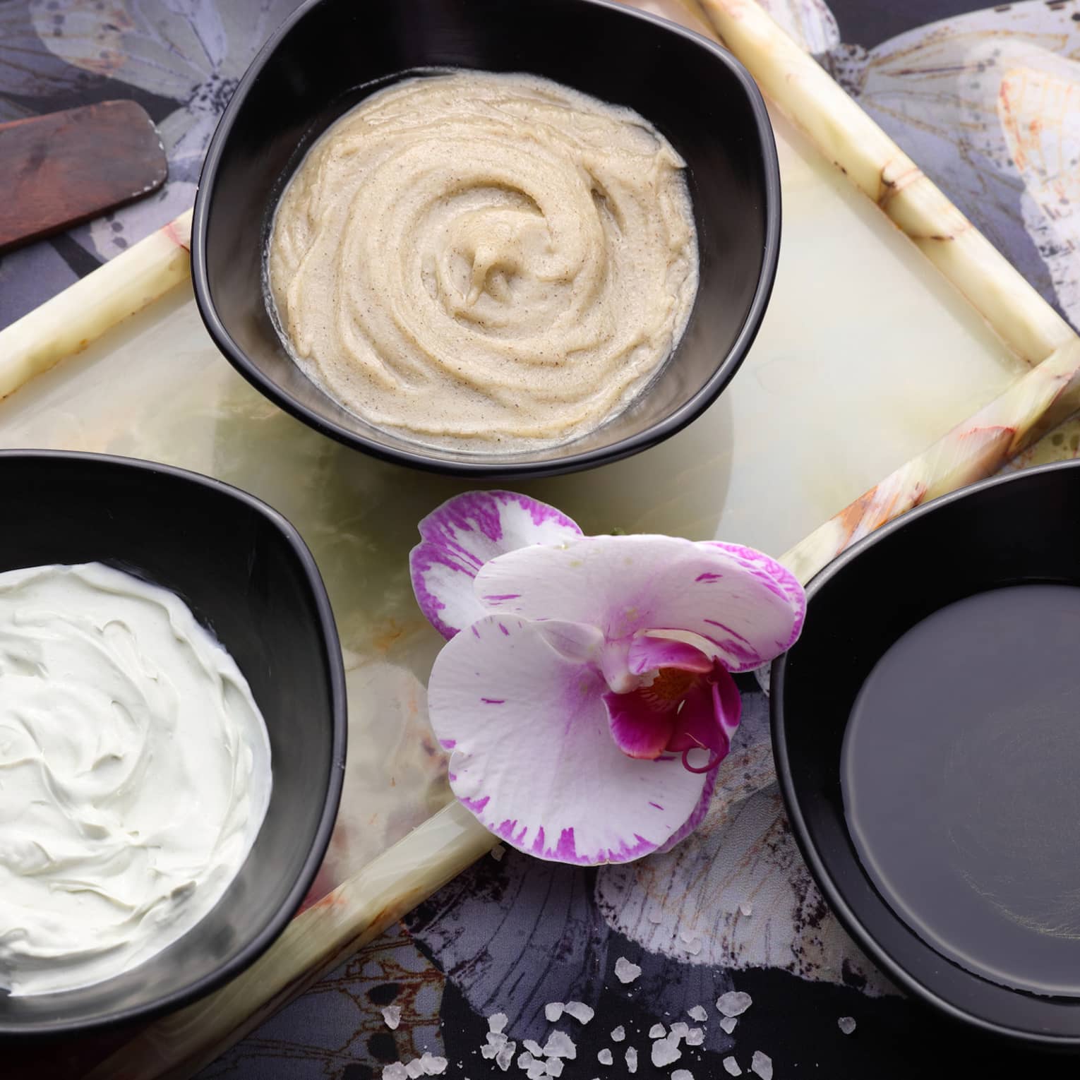 Looking down at three small black bowls with lotions, oils, fresh orchid flower