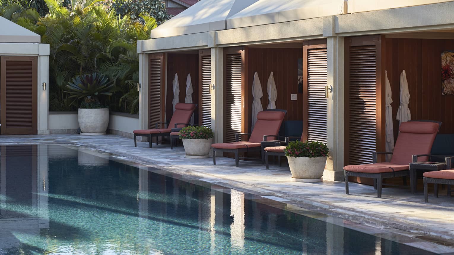 Cabanas line outdoor swimming pool deck under palm trees and sunny sky