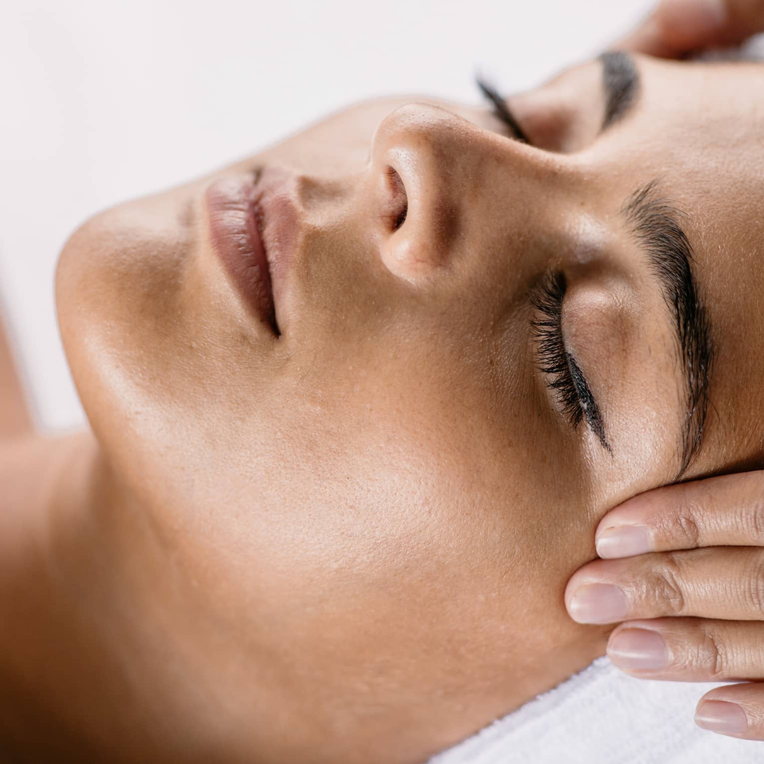 Hands massage woman's temples as she closes her eyes, spa towel around hair
