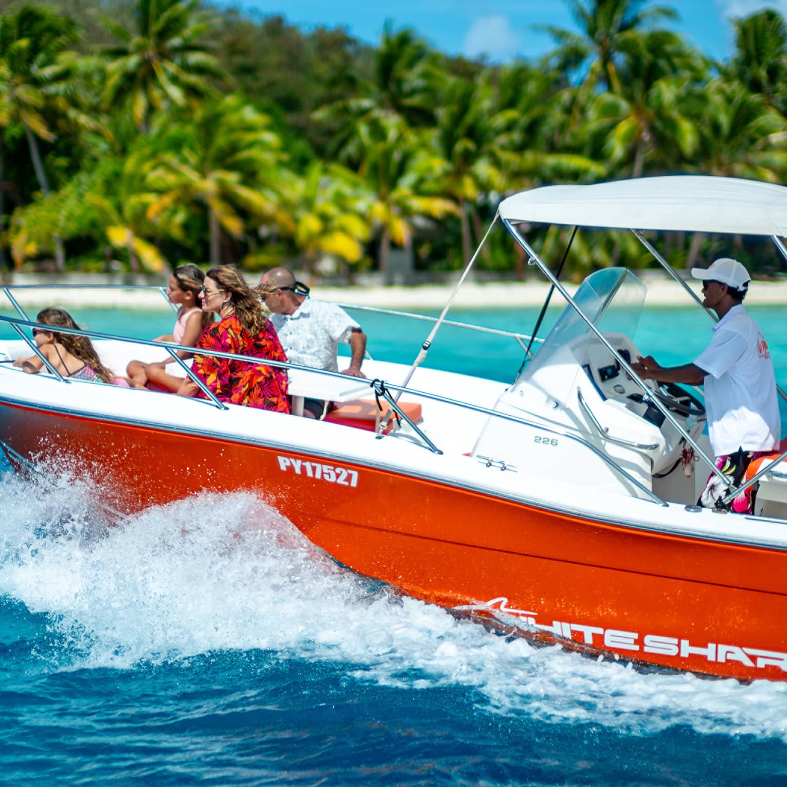 A motorboat speeds through the waves towards a palm-lined beach, a group of guests perched on the bow in the sunshine.